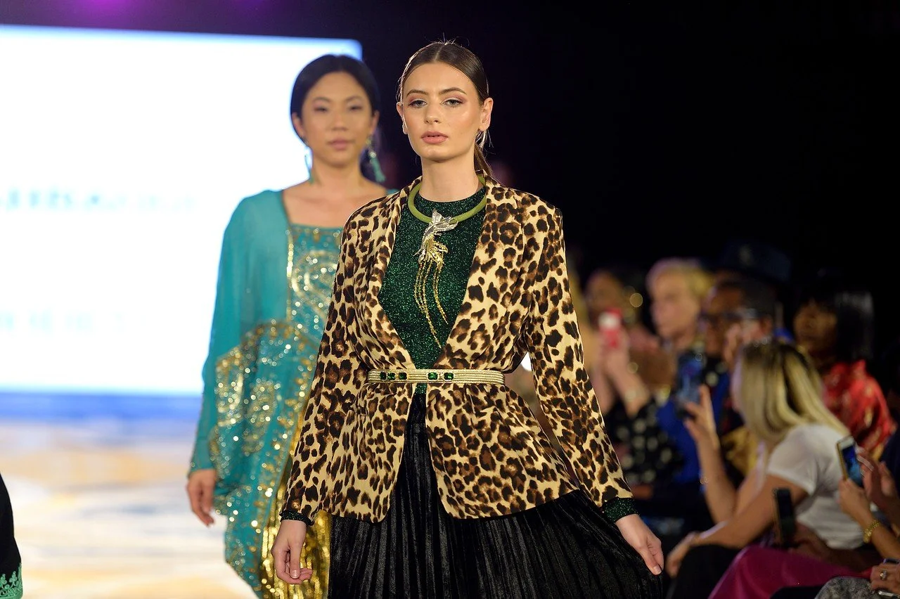 Models walking down a runway at a fashion show, with one in a leopard print blazer and green top, and the other in a turquoise dress with sequins.