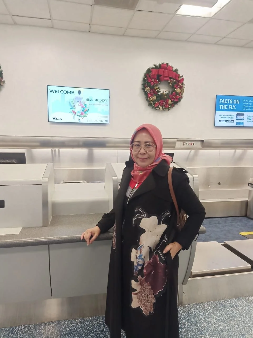 A woman wearing glasses and a pink headscarf stands at a baggage check-in counter at an airport, with a Christmas wreath hanging on the wall behind her.