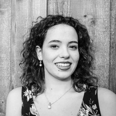 Young woman with curly hair smiling, wearing a floral dress and jewelry, standing in front of a wooden wall.