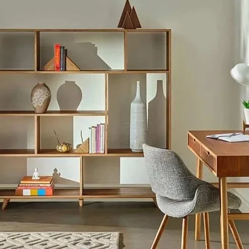 A room with a wooden bookshelf displaying decorative vases, books, and ornaments. There is a gray upholstered chair and a wooden desk with an open book and a lamp. A patterned rug is on the floor.
