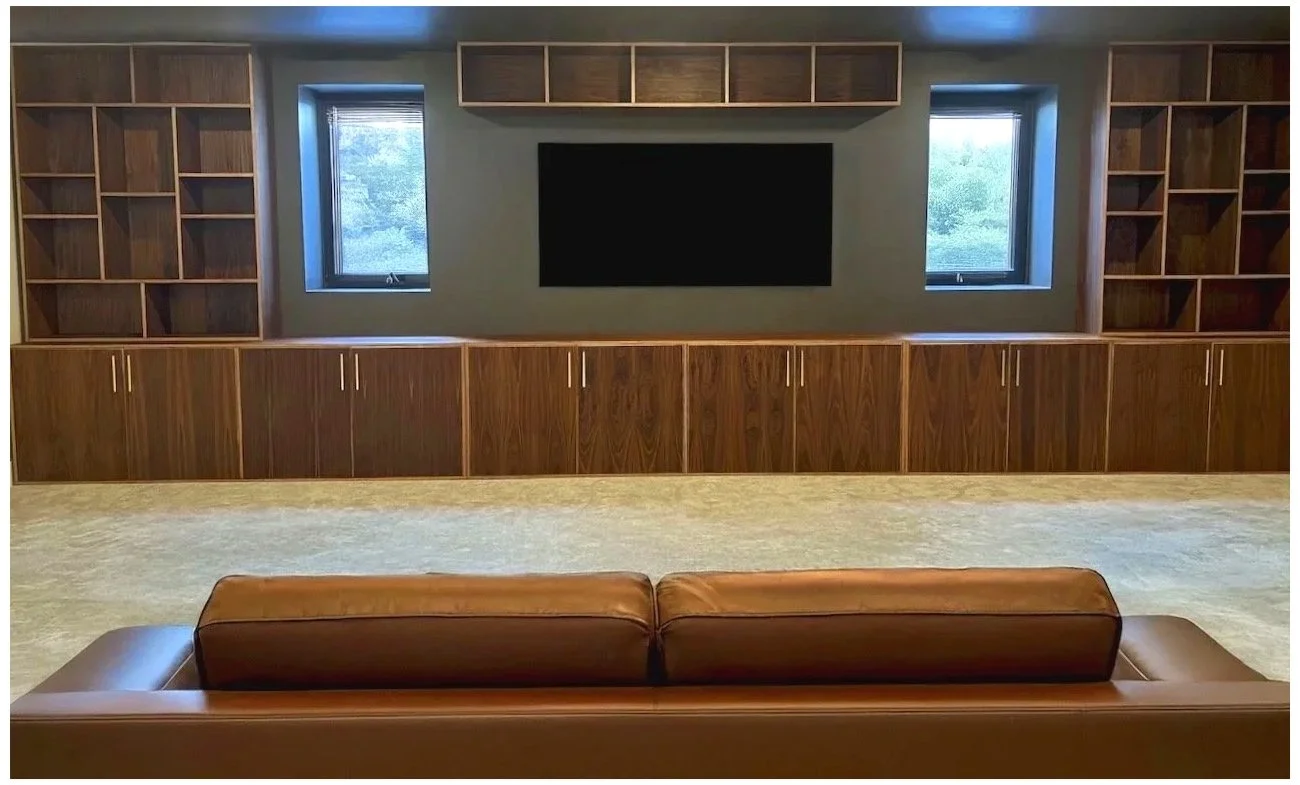 Living room with wooden built-in shelves, cabinets, and a large flat-screen TV on the wall. Two windows are on either side of the TV, allowing natural light. There's a brown leather couch in the foreground.