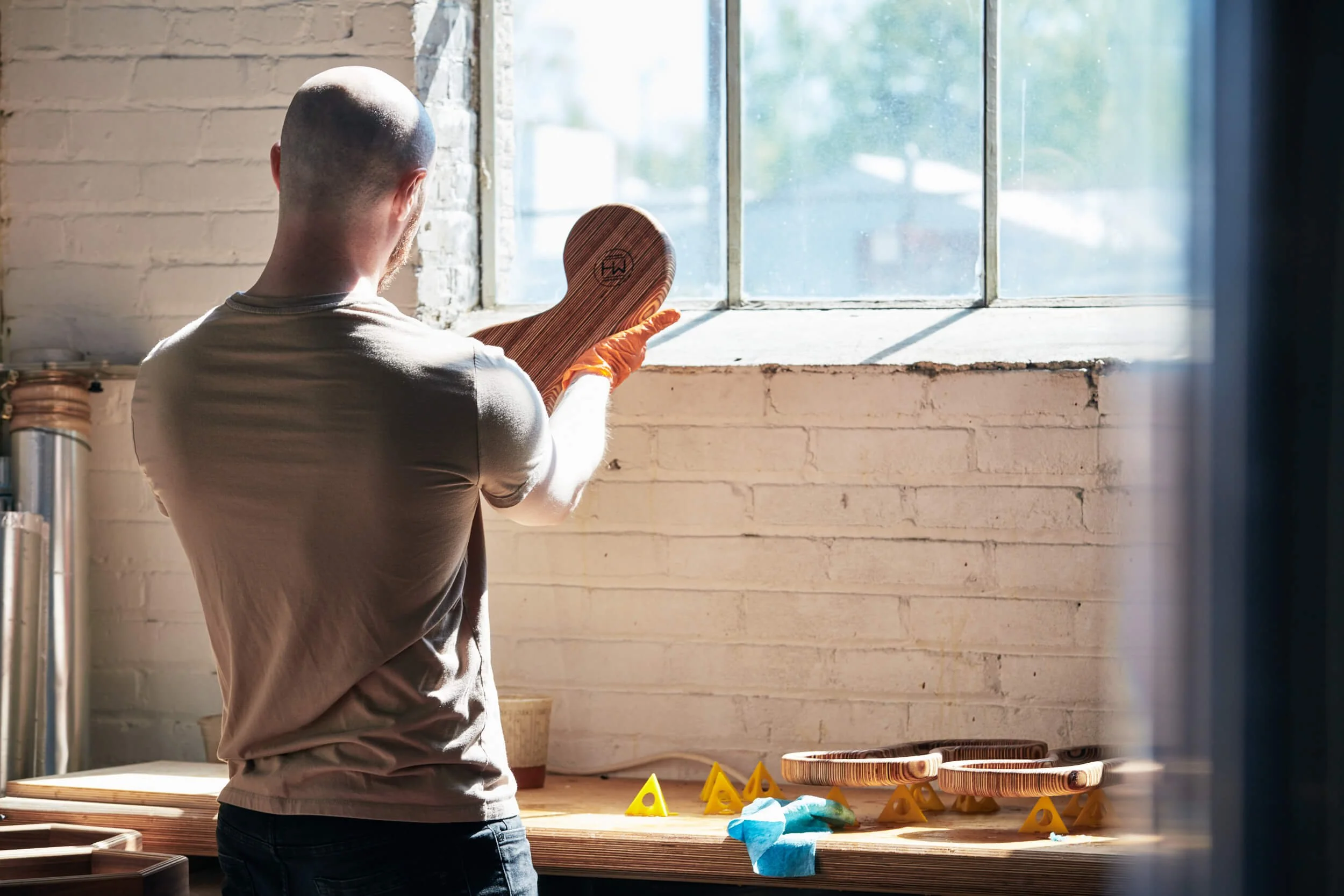 A man wearing a gray t-shirt and orange gloves using a wooden tool on a workbench in a workshop with sunlight coming through the window.