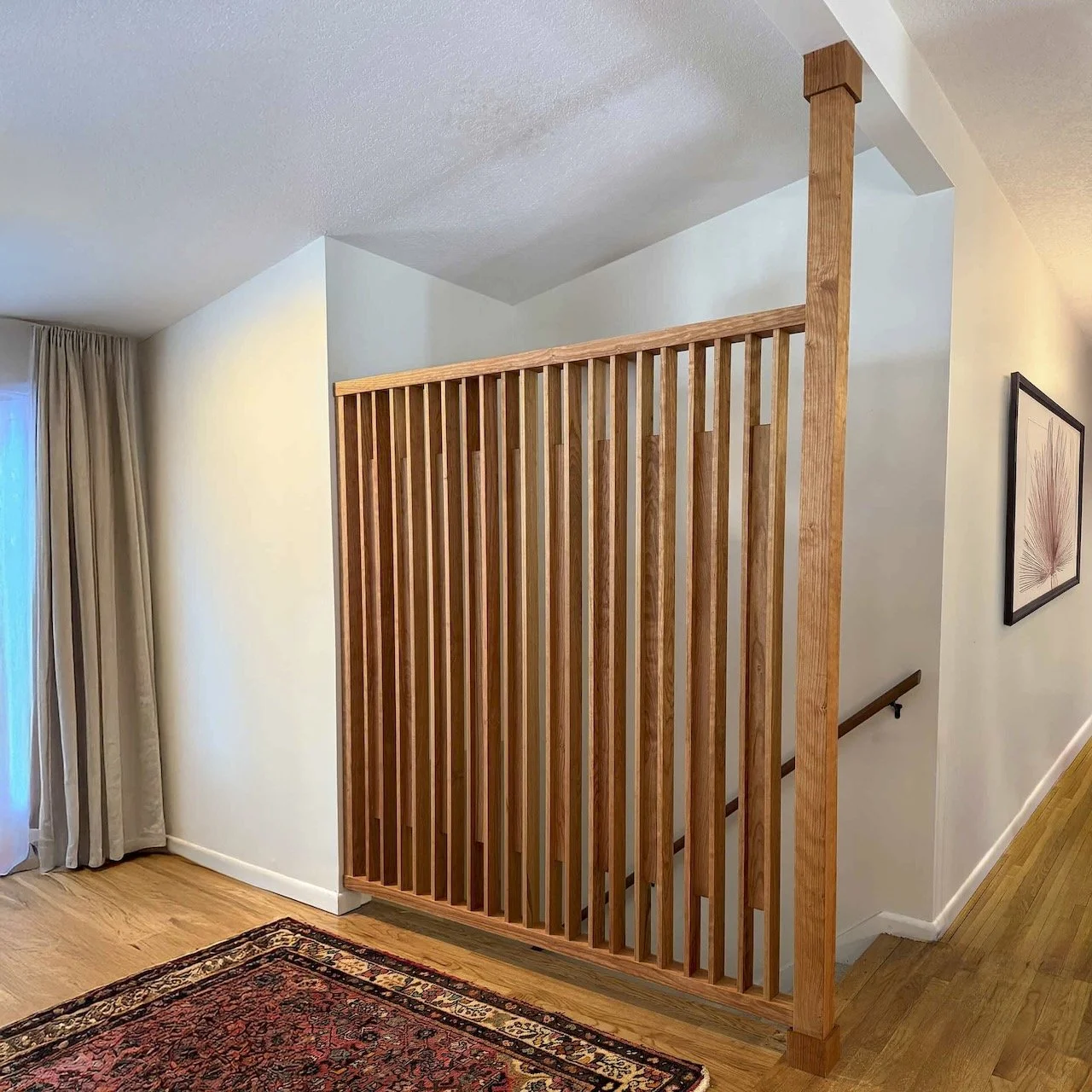 Interior view of a home staircase with a wooden slat railing and hardwood flooring, next to a wall with a framed artwork, near a window with curtains.