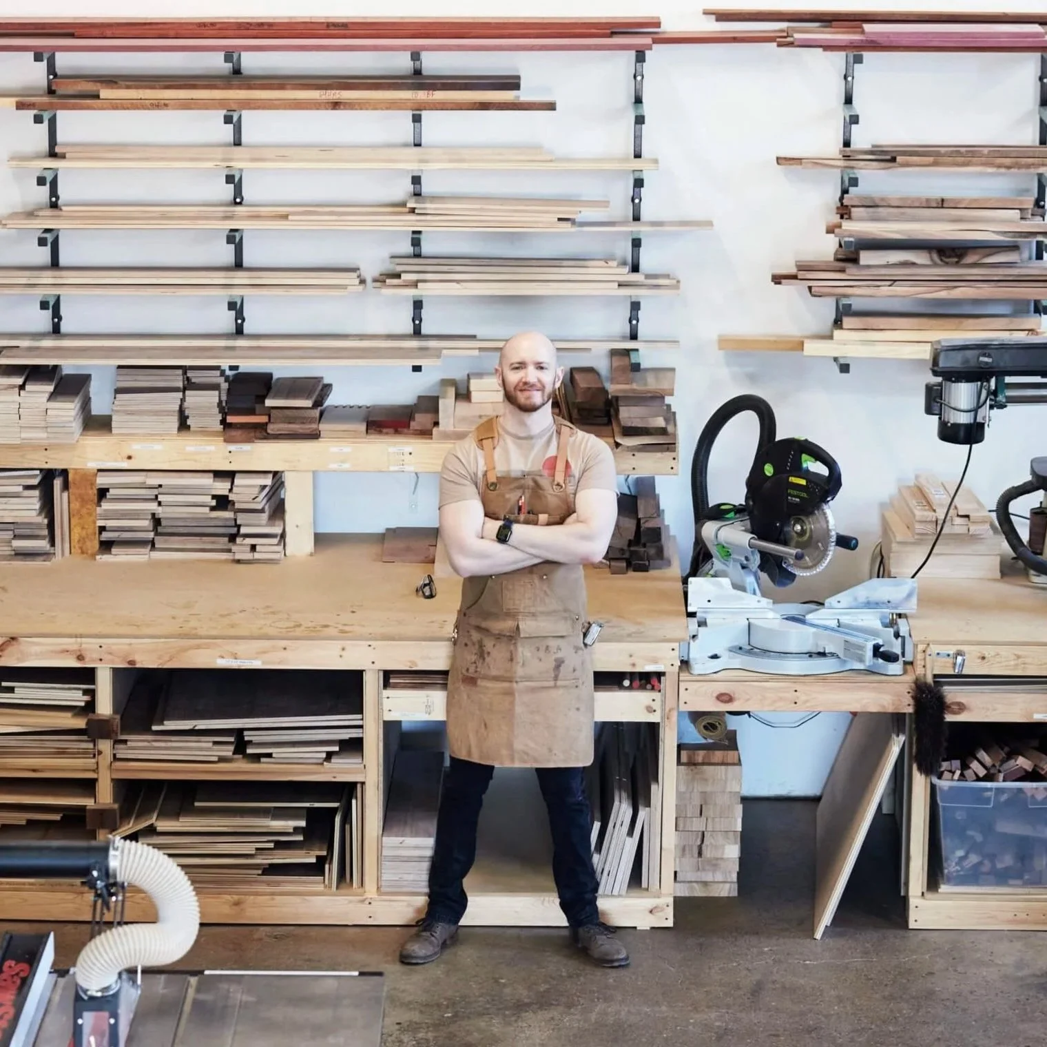 A man standing with arms crossed in a woodworking shop, surrounded by shelves and stacks of wood, with woodworking tools and equipment including a circular saw.