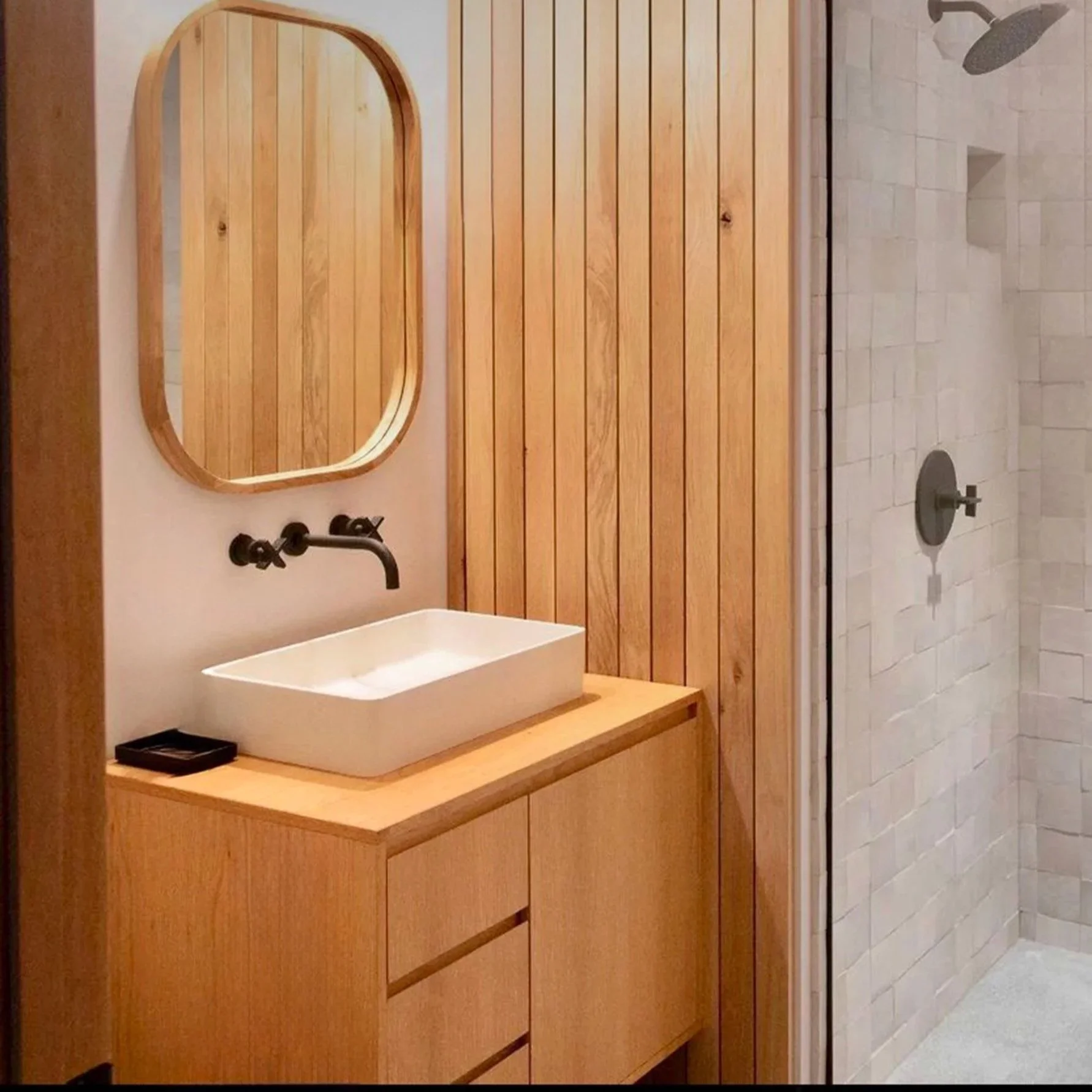 Modern bathroom with wooden wall panels, a white rectangular vessel sink, black wall-mounted faucet, mirror, small black tray, and a separate shower area with beige tiled walls and a rainfall showerhead.