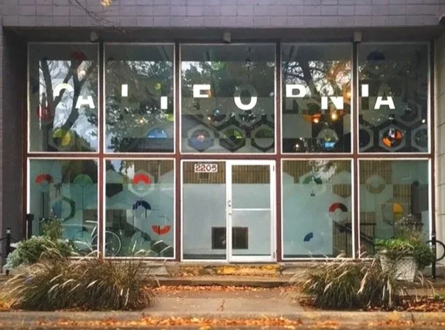 The storefront has large glass windows with the letters 'H I G H C O R N I A' displayed across the top. Inside, various objects including bicycles and colorful geometric shapes are visible. Outside, there are ornamental grasses and plants near the entrance.