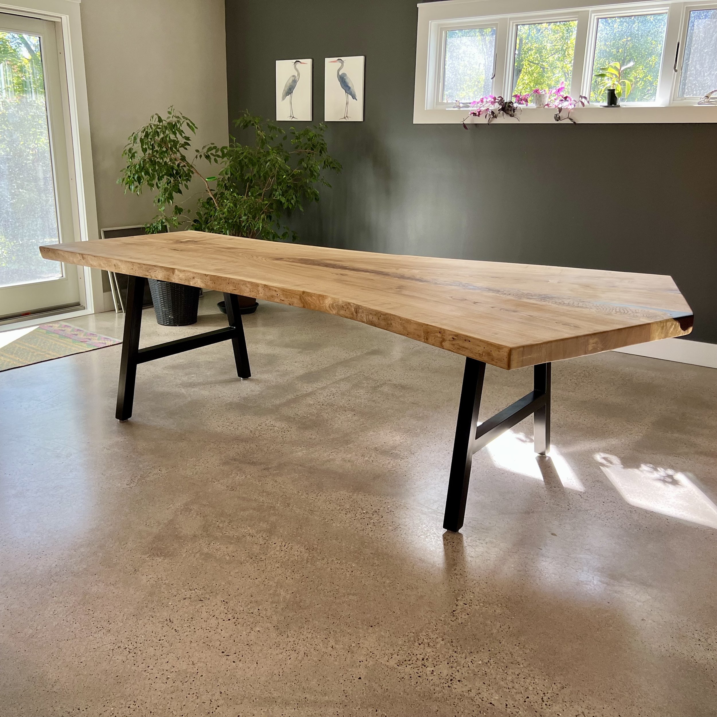 A modern wooden dining table with black legs in a room with beige flooring, green walls, potted plants, and framed bird illustrations on the wall.