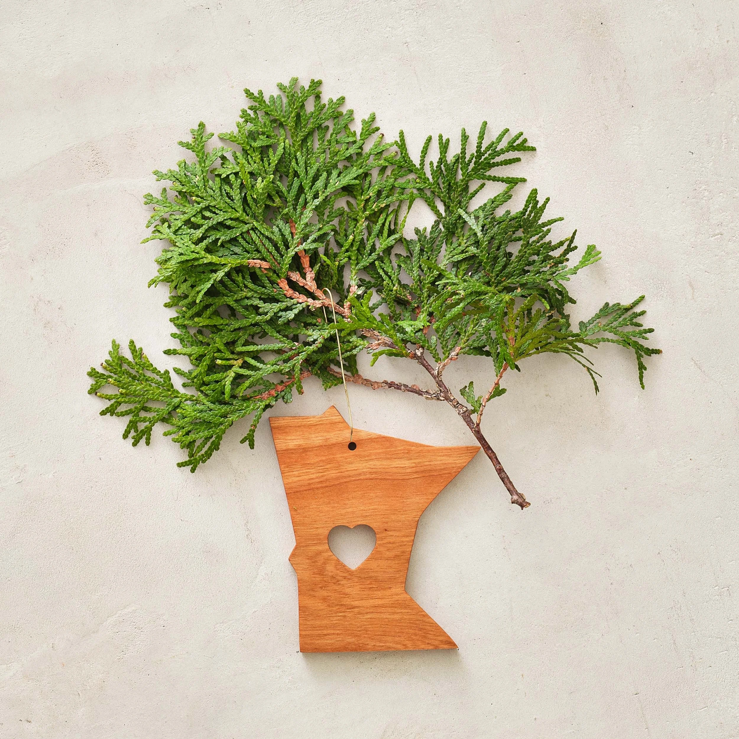 A sprig of green foliage attached to a wooden cutout of the state of Minnesota with a heart-shaped cutout in the center, hanging by a string.