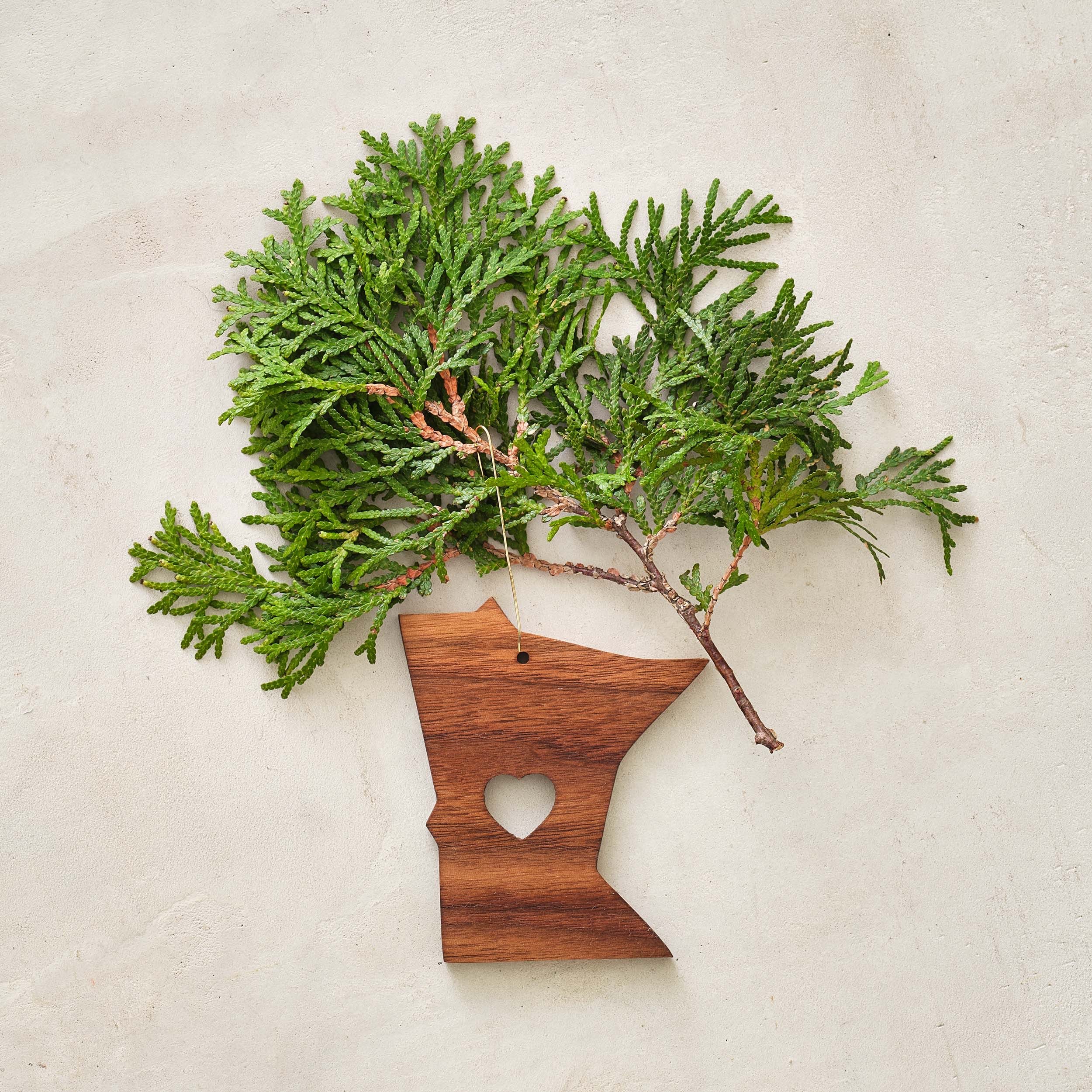 A wooden ornament in the shape of Minnesota with a heart cutout in the center, with green cedar branches attached, on a light-colored surface.