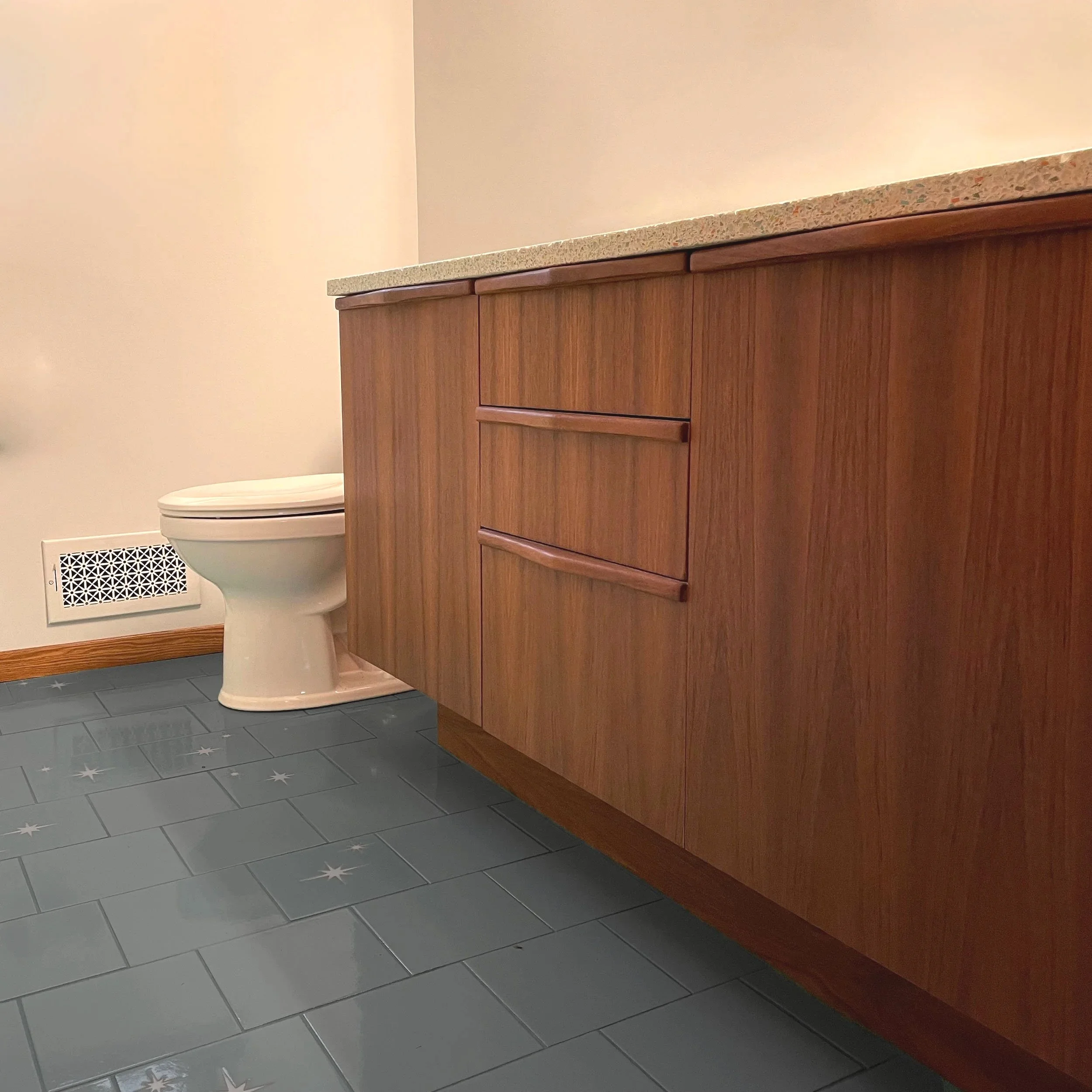 A bathroom with a toilet and a wooden cabinet with a granite countertop, gray tiled floor with star patterns, and a white wall with a heating vent.
