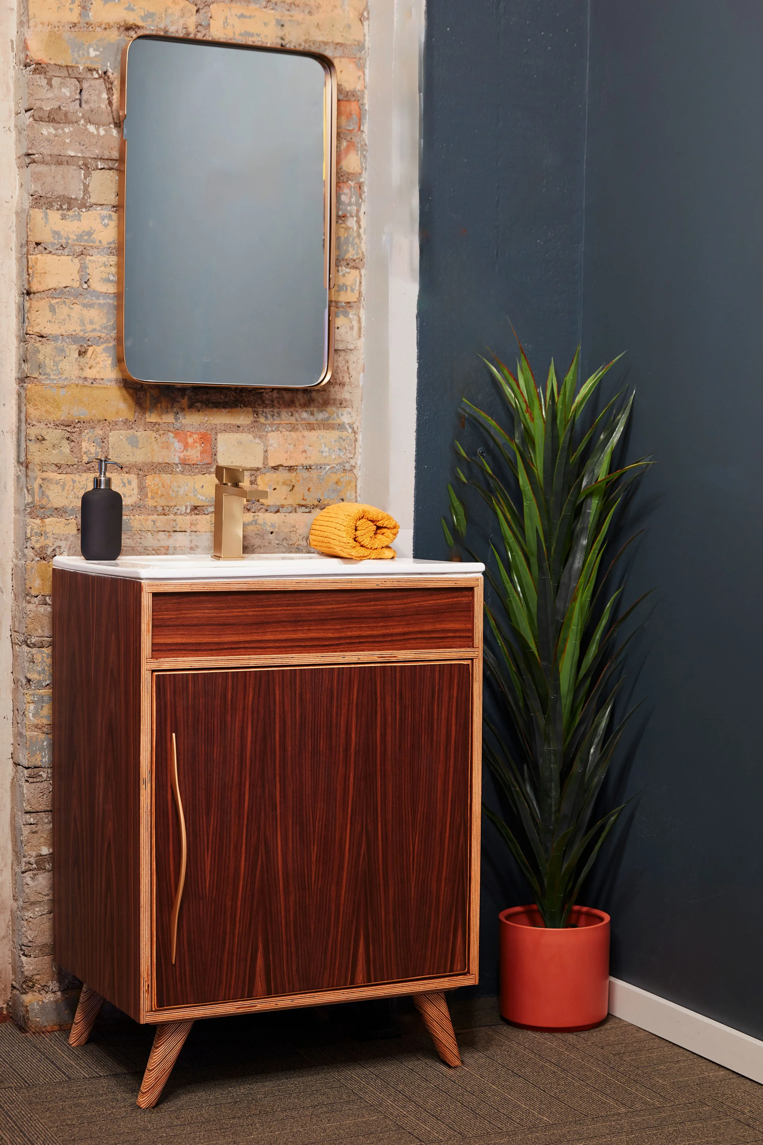 A bathroom vanity with a wooden cabinet, a white countertop, and a gold faucet, a black soap dispenser, a yellow rolled towel, and a large green plant in a red pot, with a mirror on a brick wall and dark painted wall behind.