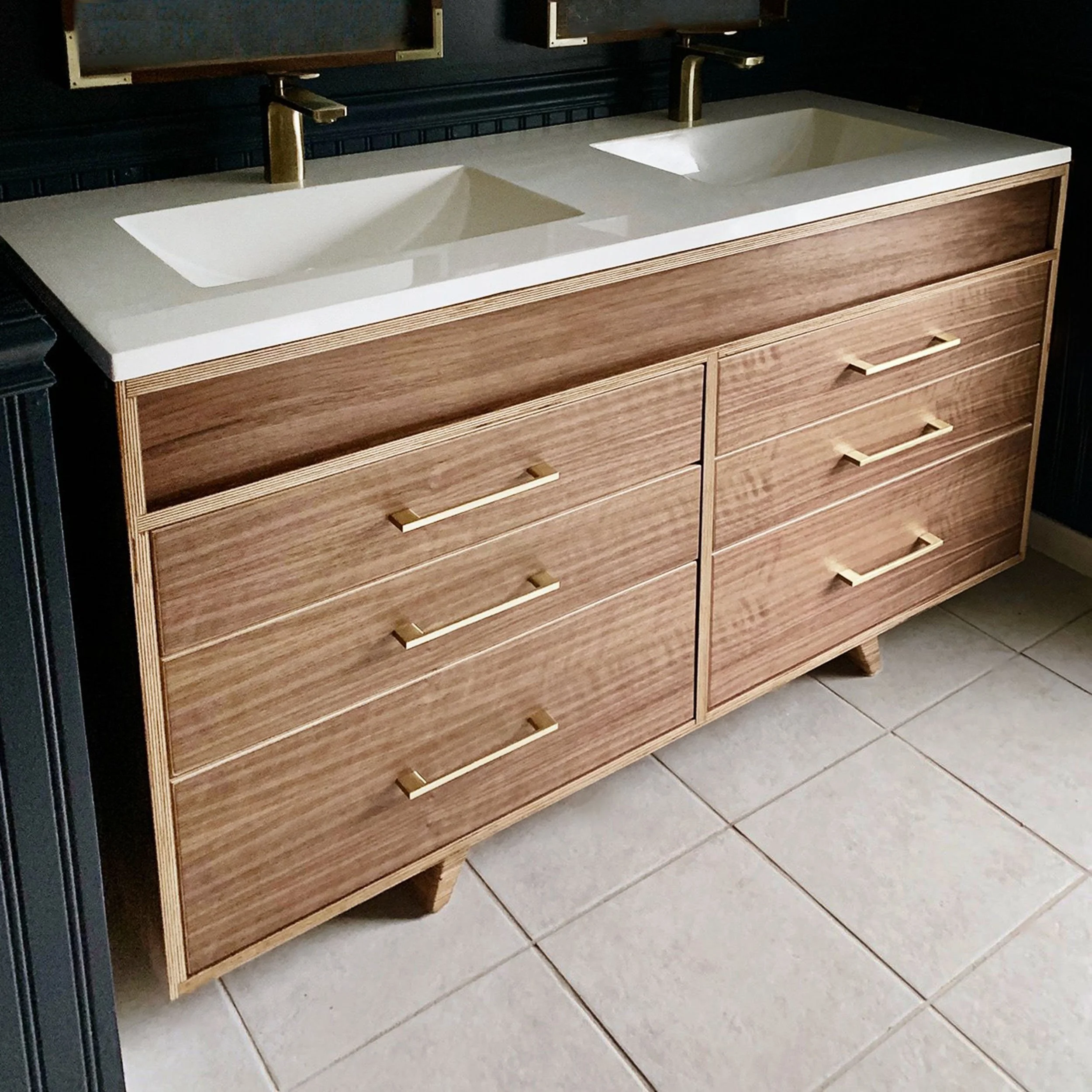 Wooden double vanity with two sinks, white countertops, and gold handles, situated on tiled floor.