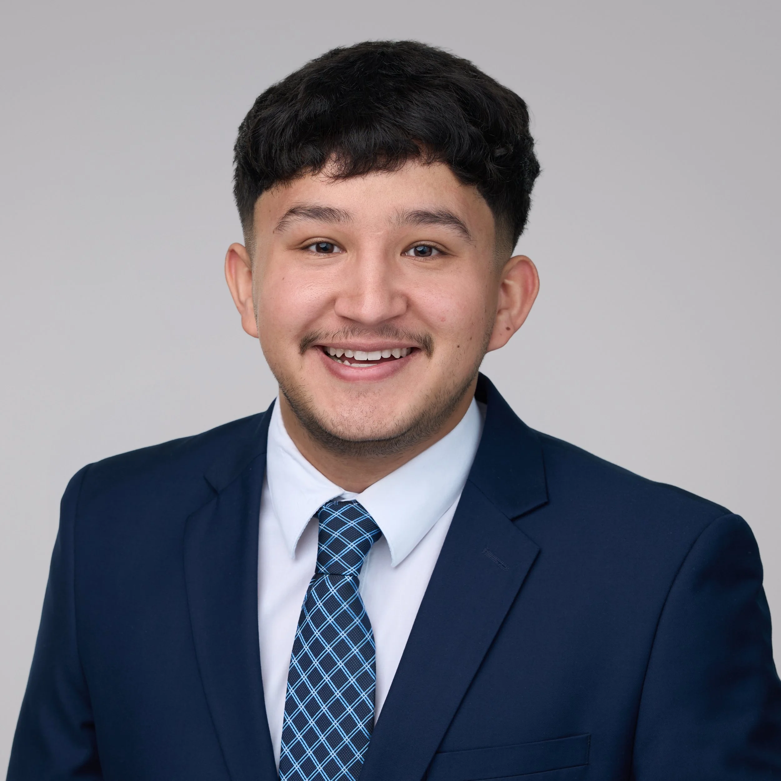 A smiling young man with dark hair, wearing a navy blue suit, white shirt, and a blue patterned tie, against a plain light gray background.