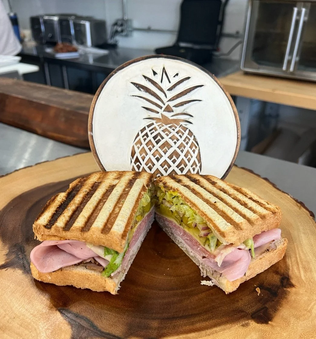 A grilled sandwich with ham, lettuce, and cheese is cut in half and placed on a wooden board. Behind it is a decorative pineapple design.