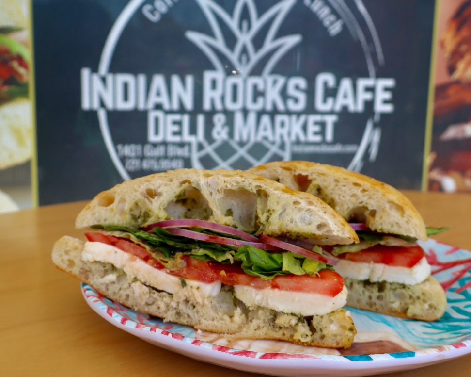 A grilled sandwich with ham, lettuce, and cheese is cut in half and placed on a wooden board. Behind it is a decorative pineapple design.