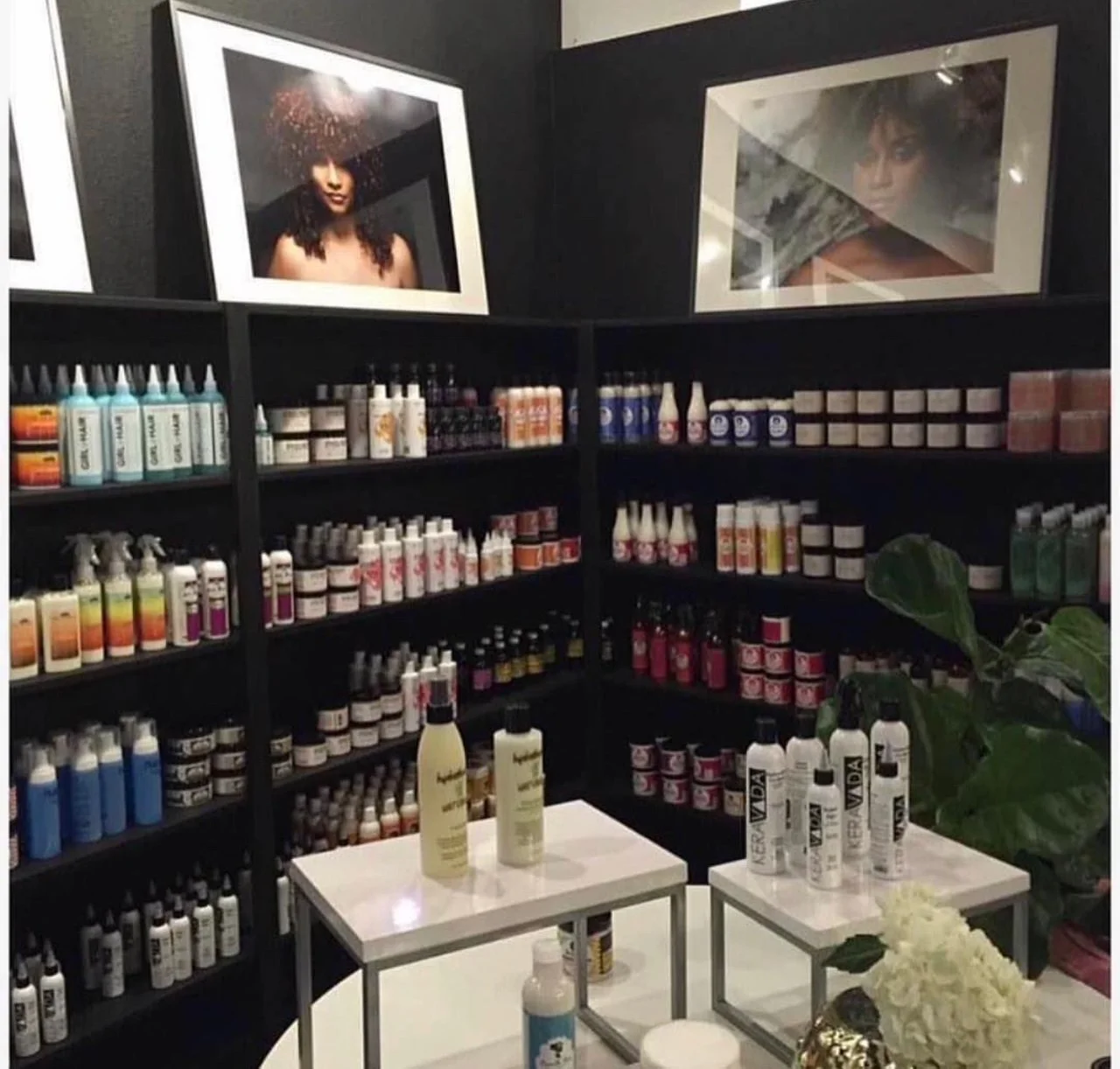 A display of various hair care and styling products on black shelves, with two framed portraits of women above the shelves, and a table with bottles and a flower arrangement in the foreground.