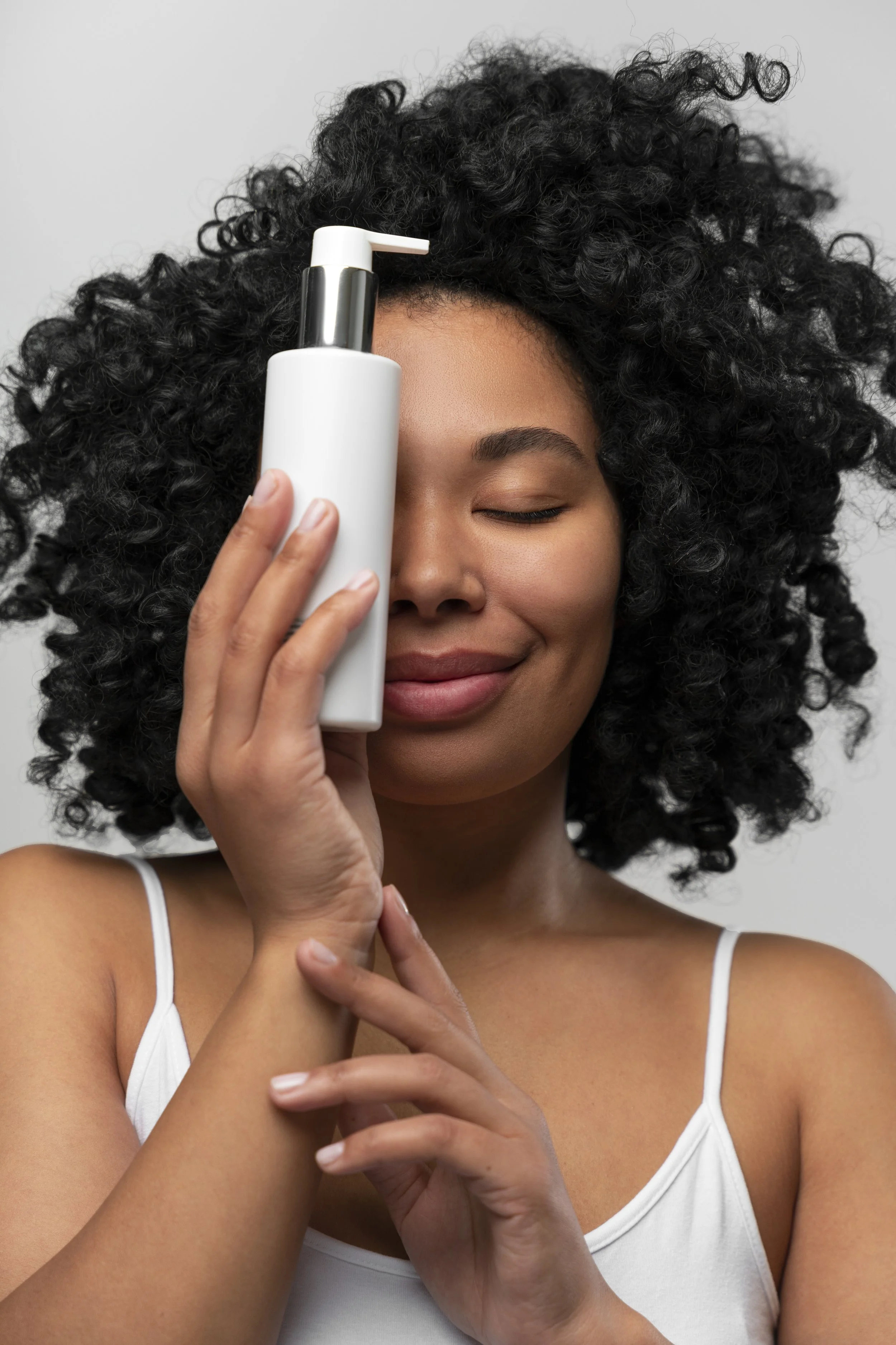 A woman with curly black hair holding a white haircare pump bottle against her face with her eyes closed, smiling gently.