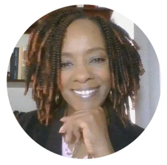 A person with braided hair smiling, wearing a dark-colored blazer, with a bookshelf in the background.