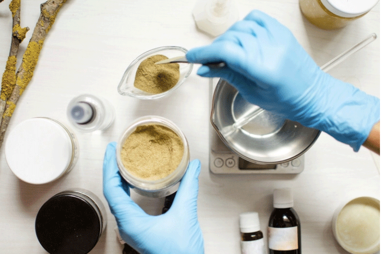 Gloved hands scooping a brown powder into a container, surrounded by bottles and jars on a white surface, with a scale.