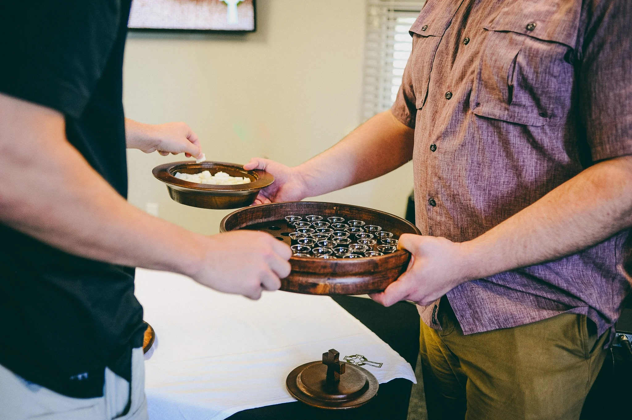Two people exchanging communion elements during a religious ceremony, one holding a tray with small glasses and the other holding a plate of bread.