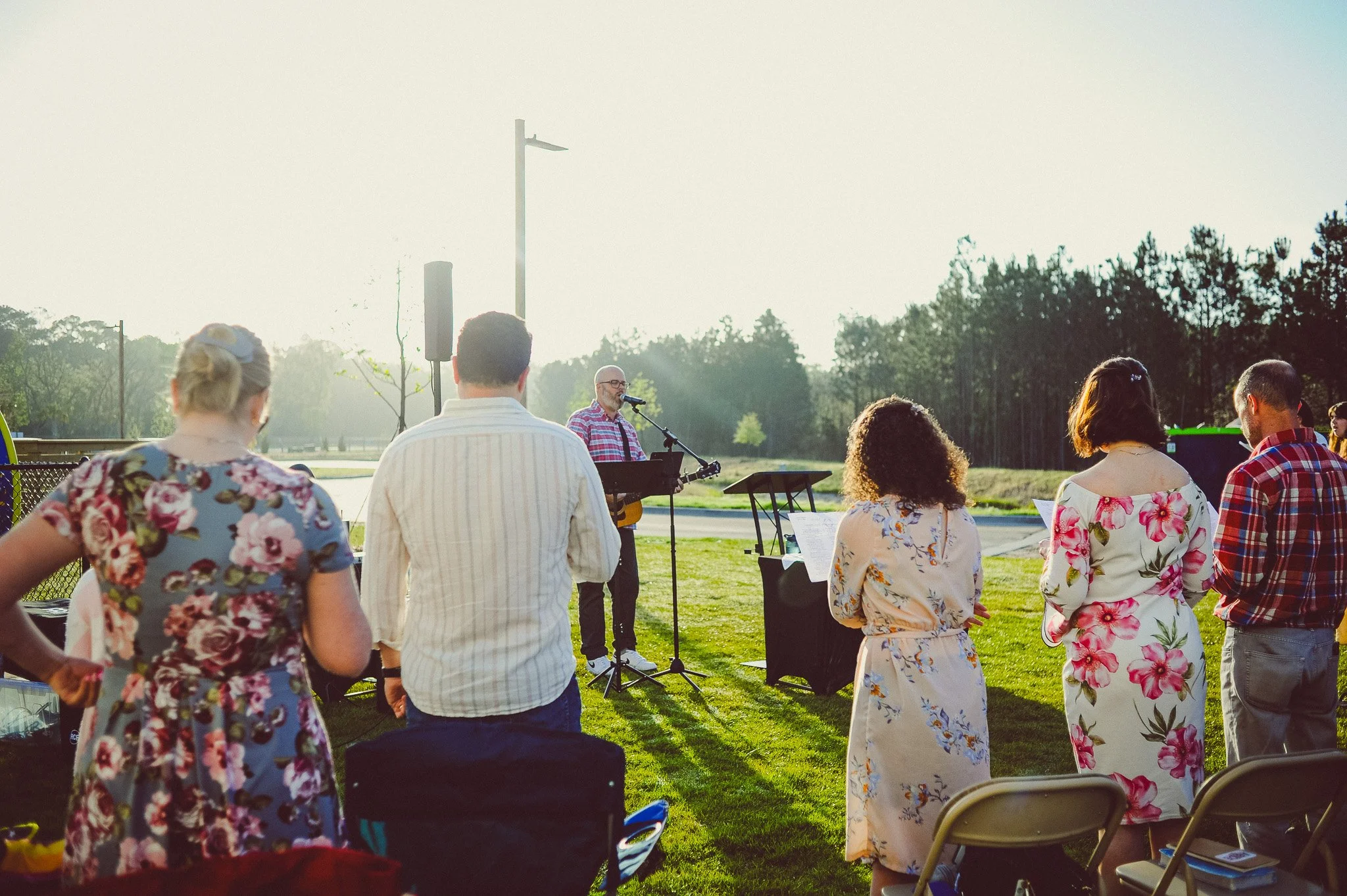 People gathered outdoors at a wedding or event during sunset, facing a man singing and playing guitar, with chairs and music stands around.