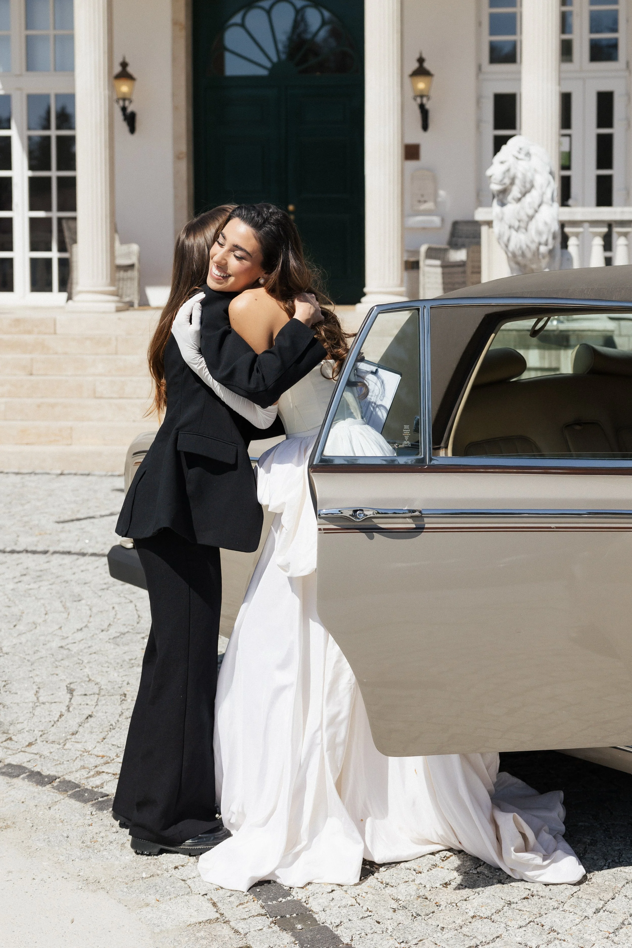 Zwei Frauen umarmen sich vor einem klassischen Auto auf einem gepflasterten Platz, im Hintergrund eine große Hausfassade mit Säulen und einer Statue eines Löwen.