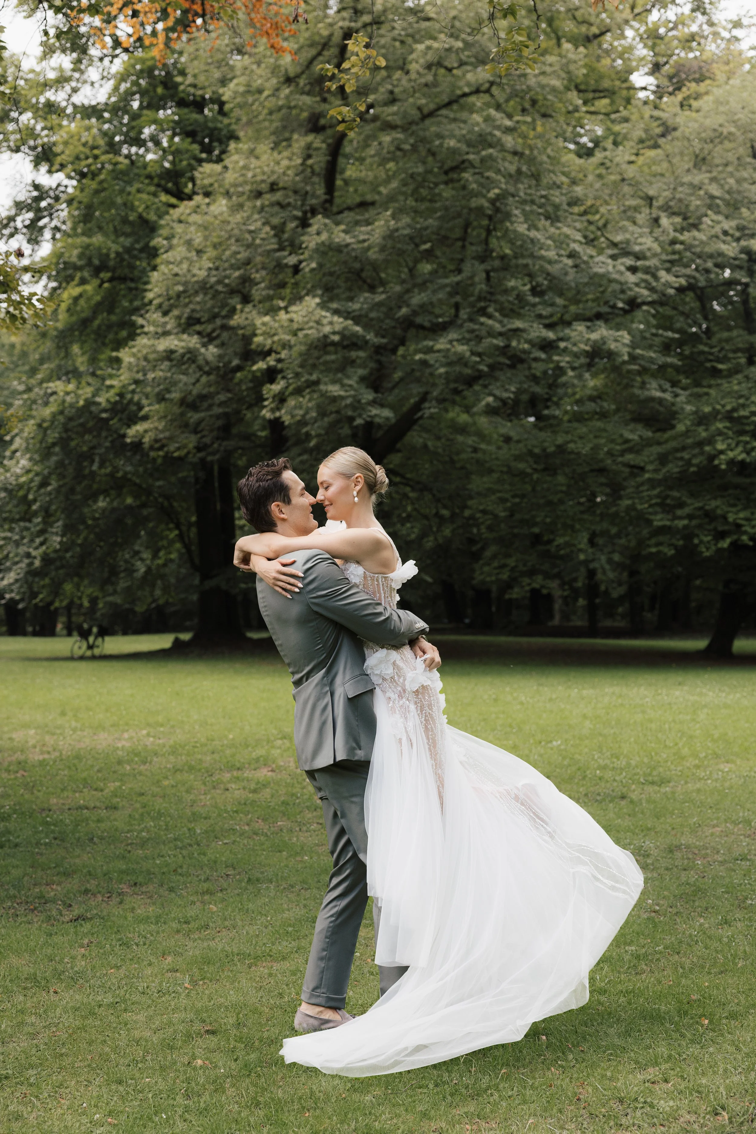 Ein Brautpaar in Hochzeitskleidung umarmt sich in einem Park mit grünen Bäumen im Hintergrund.