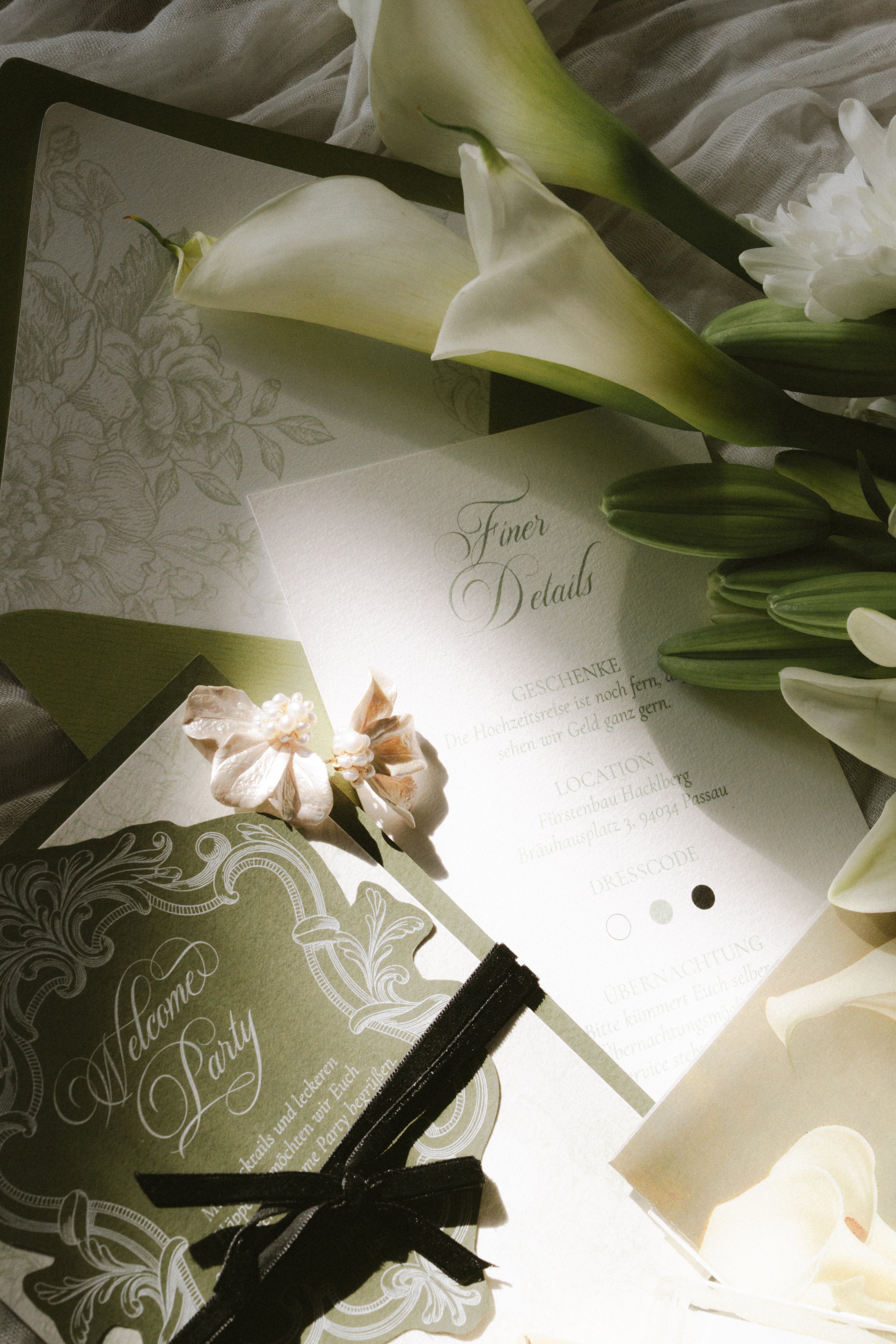 Dekorative Hochzeitseinladungen mit Blumenmotiven und kalligraphischem Text auf einem Stoffhintergrund, umgeben von weißen Lilienblumen.