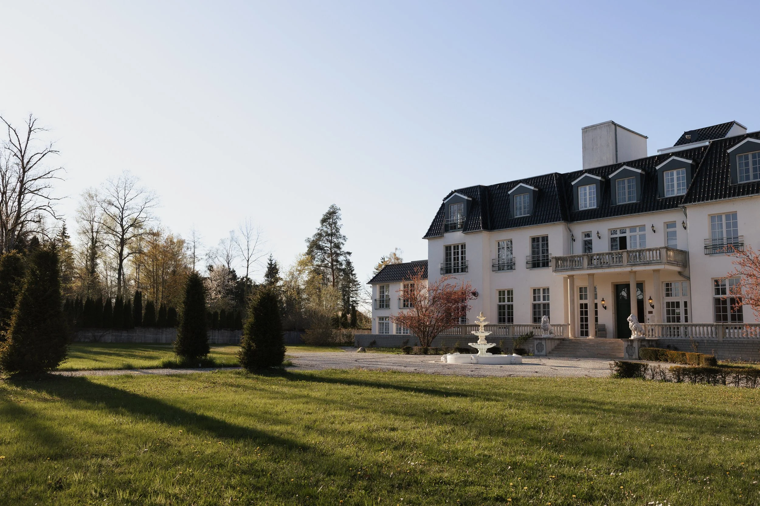 Ein großes, weißes Herrenhaus mit schwarzem Dach, mehrere Stockwerke, Balkon und Statuen vor dem Eingang, umgeben von einem gepflegten Garten mit Springbrunnen, Bäumen und Rasen bei sonnigem Himmel.
