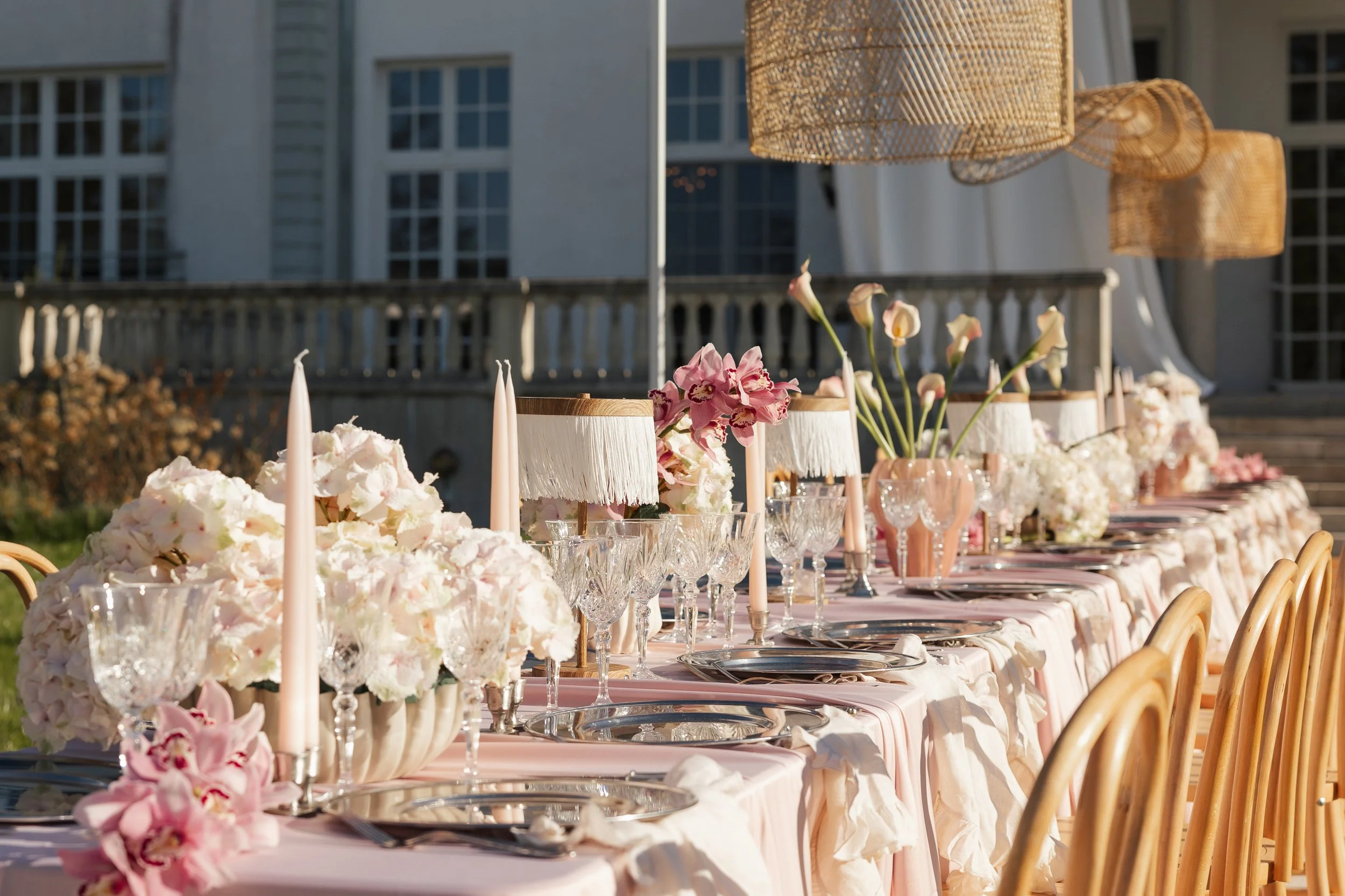 Ein schön gedeckter Esstisch im Freien mit goldfarbenen Stühlen, weißen Tischtüchern, mehreren Blumenarrangements in weißen Vasen, Kerzen und Kristallgläsern bei Sonnenuntergang vor einem großen weißen Gebäude.