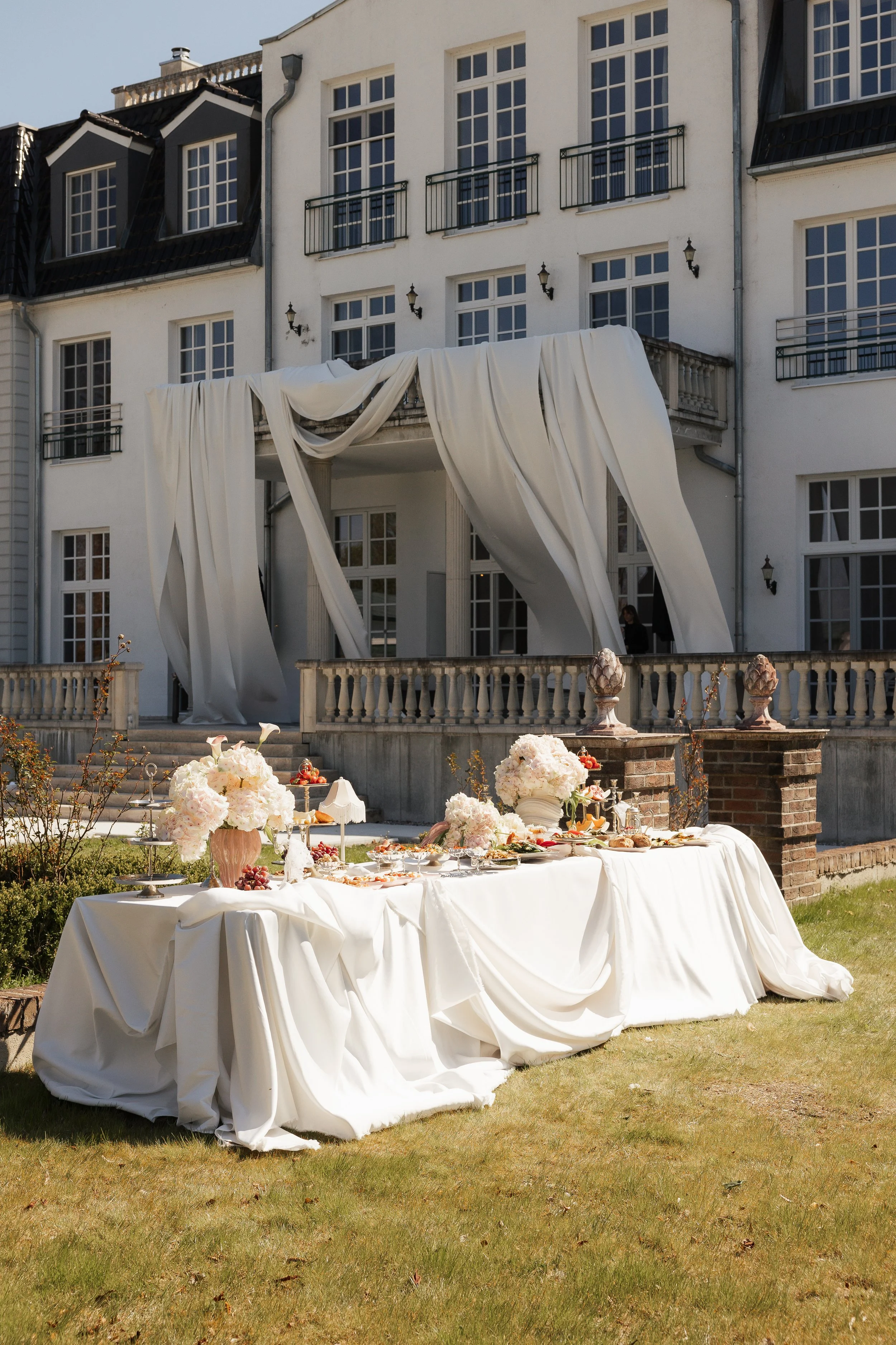 Ein Gartentisch mit weißer Tischdecke und Blumenarrangements, gedeckt für ein Fest, vor einem weißen Gebäude mit mehreren Fenstern und Balkonen, im Freien bei Tageslicht.