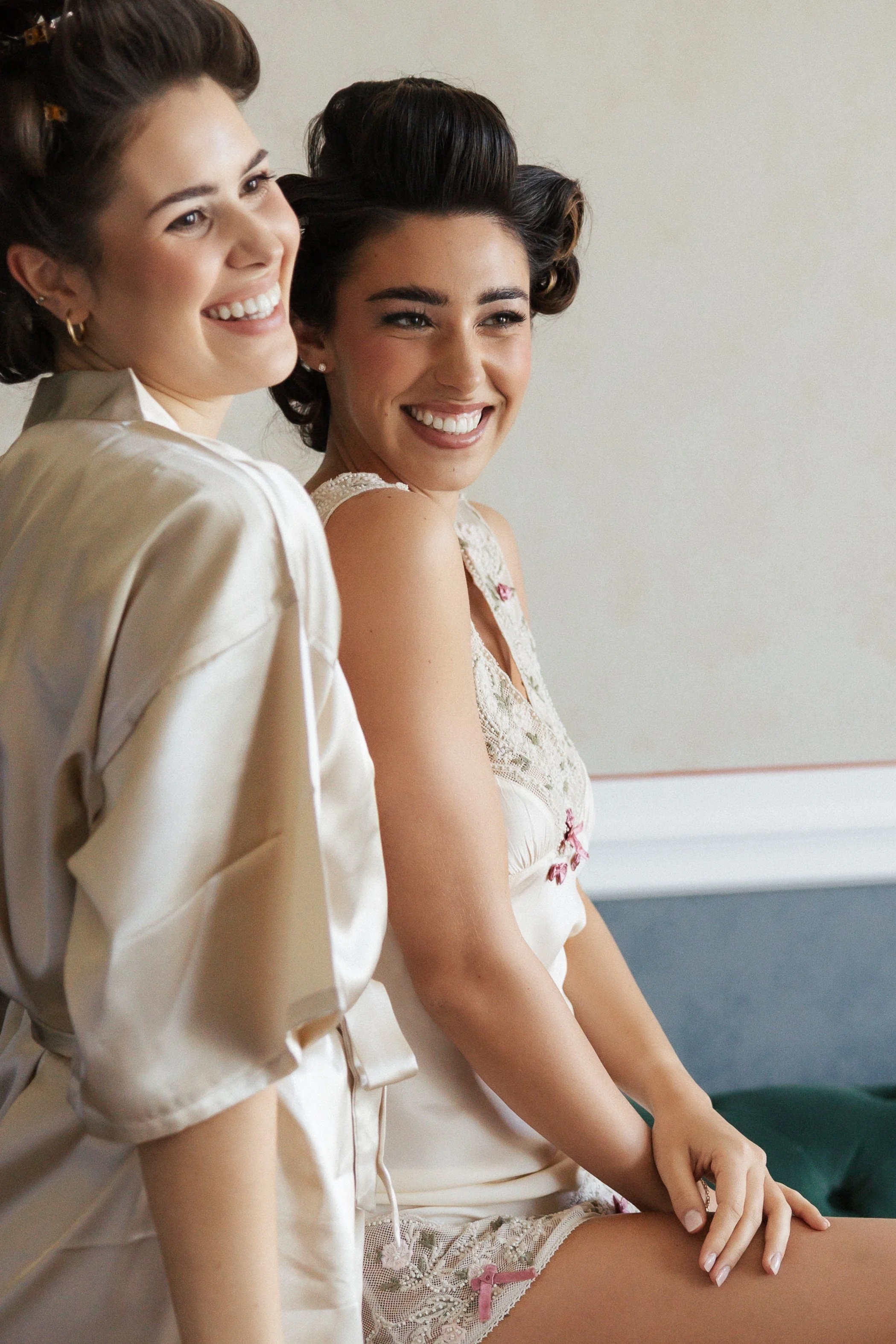 Zwei Frauen sitzen nebeneinander, lachen und tragen vintage Haarschmuck und elegante Kleidung.