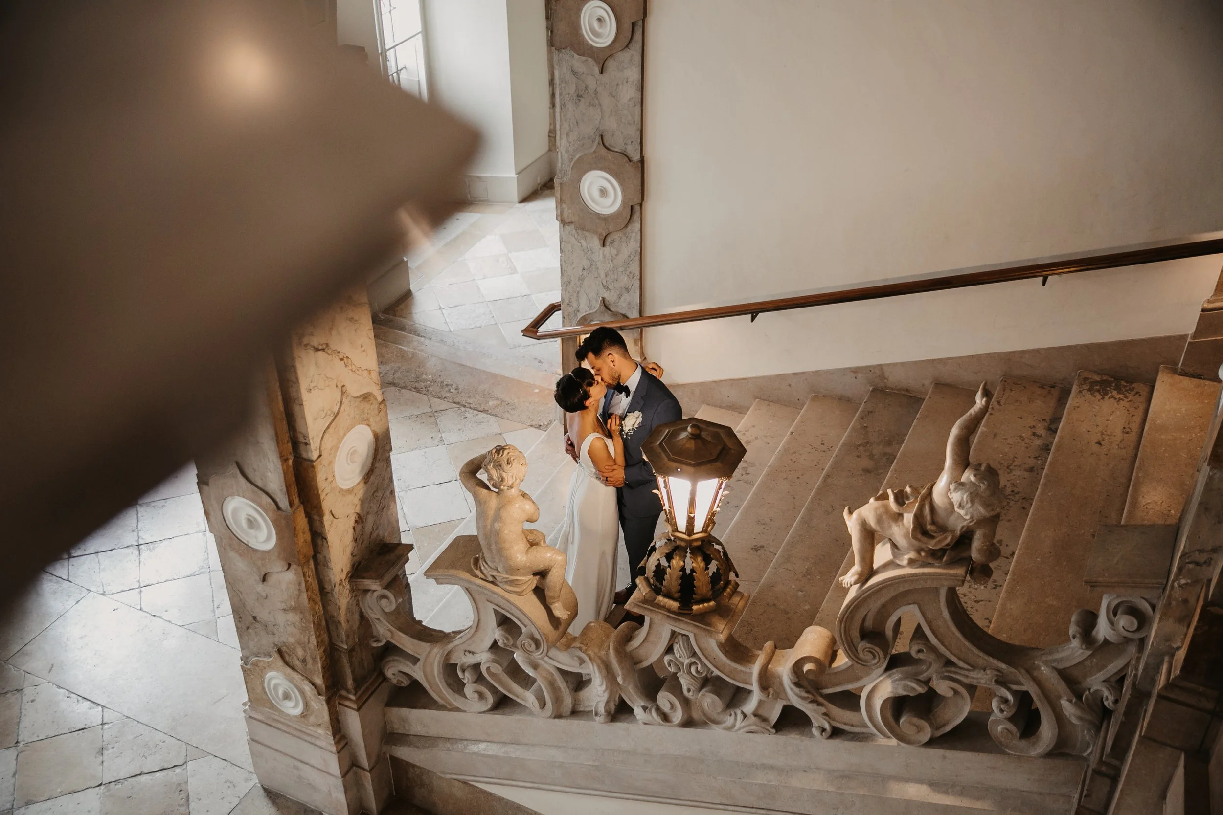 Ein Brautpaar in formeller Kleidung steht auf einer eleganten Treppe, die mit barocken Skulpturen und einem antiken Laternenlampen dekoriert ist. Die Szene ist von oben fotografiert.