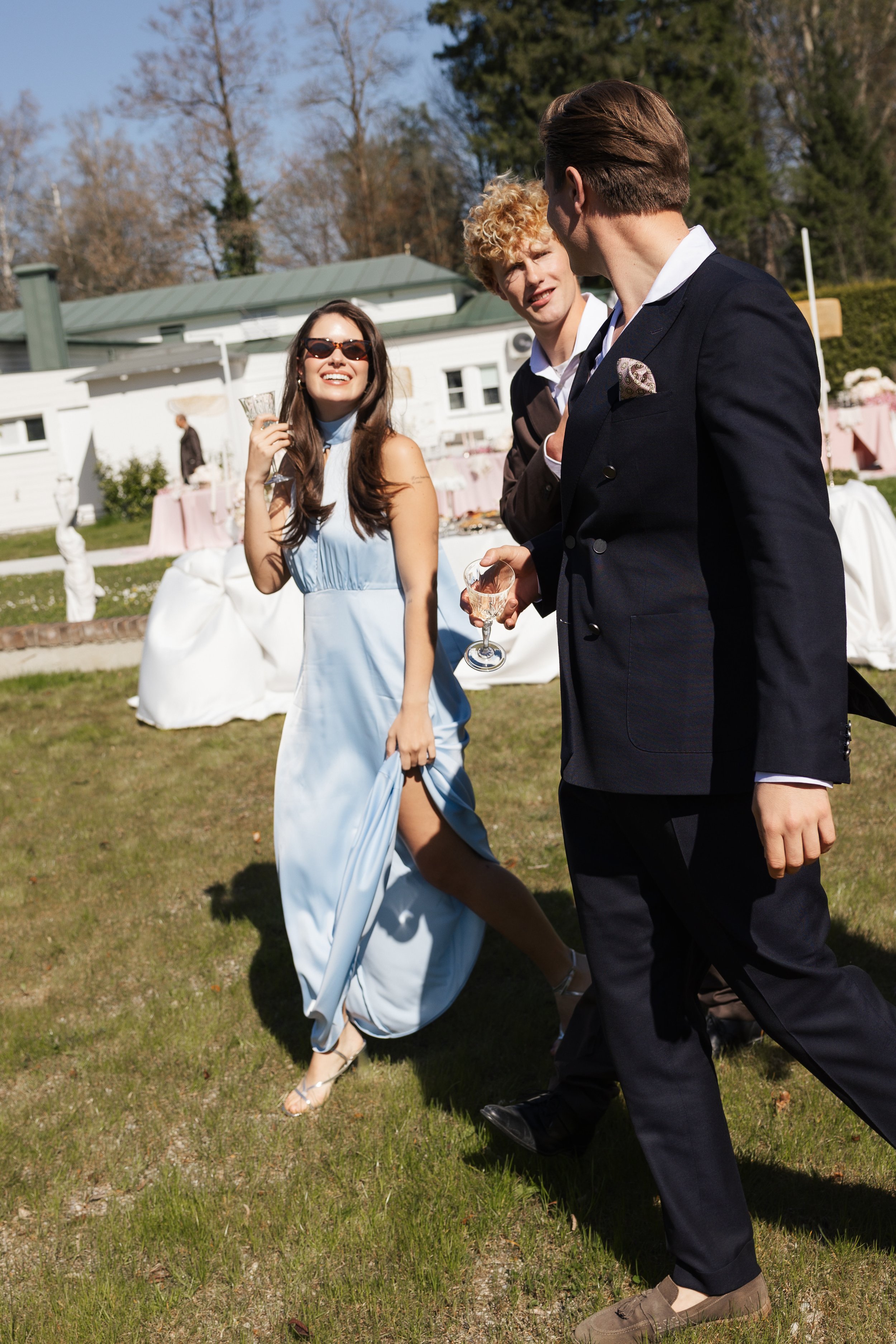 Menschen bei einer Hochzeit im Garten, eine Frau trägt ein hellblaues Kleid mit high heels, zwei Männer in Anzügen, einer mit Sonnenbrille, trinken Wein und unterhalten sich.