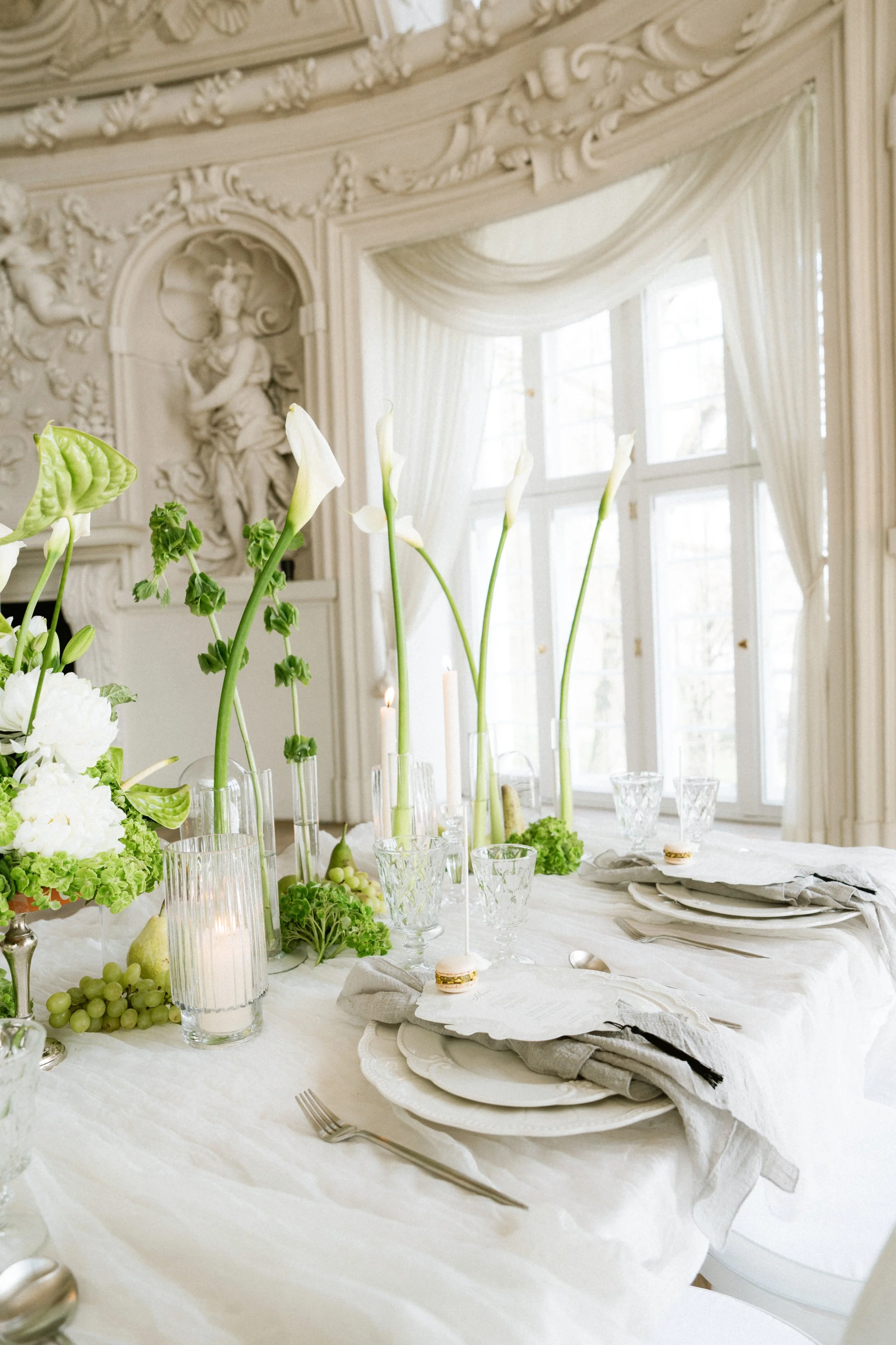 Elegante Tischdekoration in einem hellen, stilvollen Raum mit fein gedecktem Tisch. Weiße Blumen, darunter Calla-Lilien, und grüne Pflanzen schmücken die Tafel. Kristallgläser, weiße Teller und gestärkte Stoffservietten sind sorgfältig angeordnet. Im