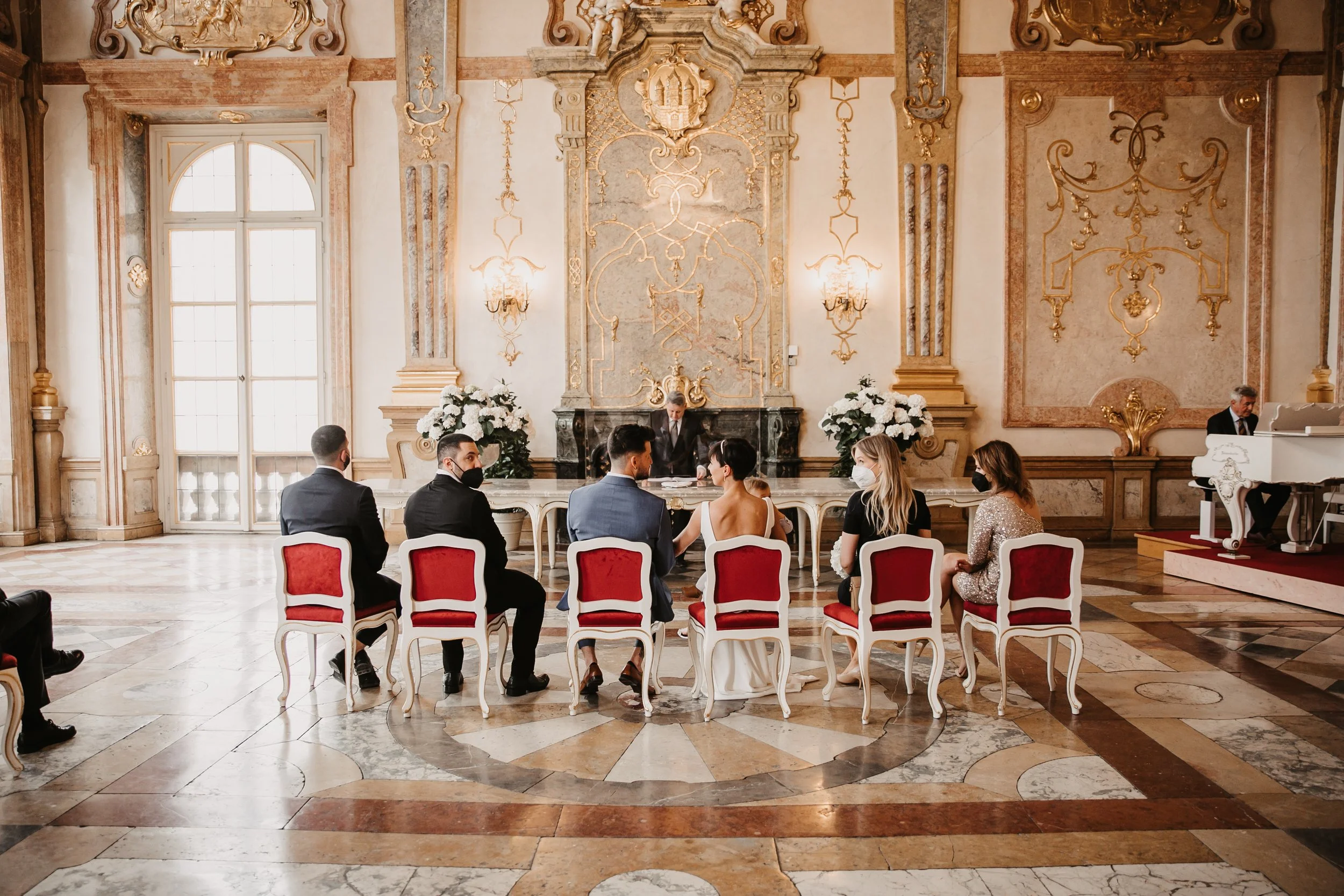 Hochzeitsszene in einem eleganten Saal mit zwölf Personen, vorne der Brautpaar bei der Trauzeremonie, pflegebedeckten Stühlen, im Hintergrund ein weißer Klavier spielender Musiker