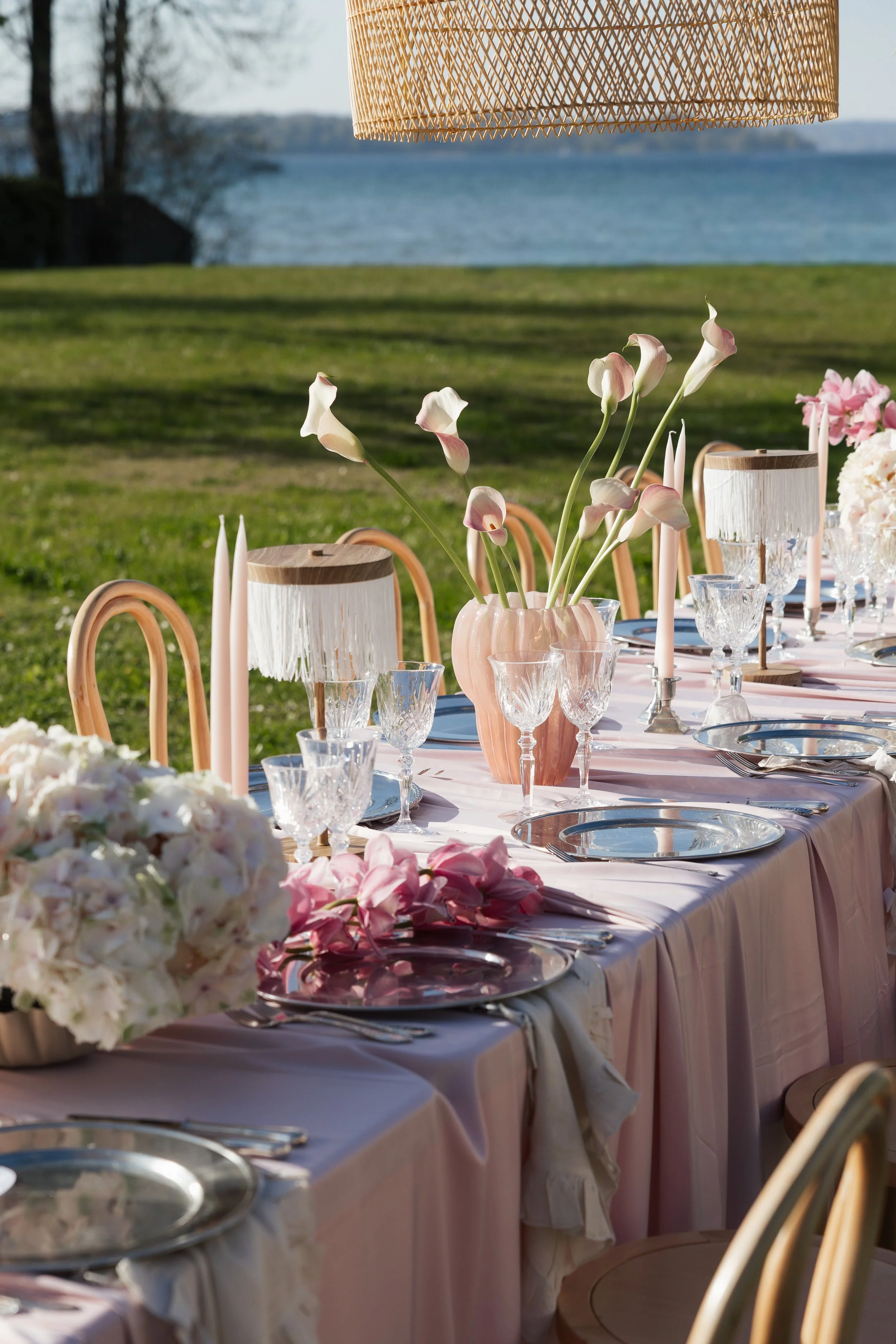Ein schön gedeckter Esstisch im Freien am Wasser, mit Blumenarrangements, Kerzen, Kristallgläsern, weißen Tellern und Besteck, unter einer rautenförmigen Lampe aus Holz.