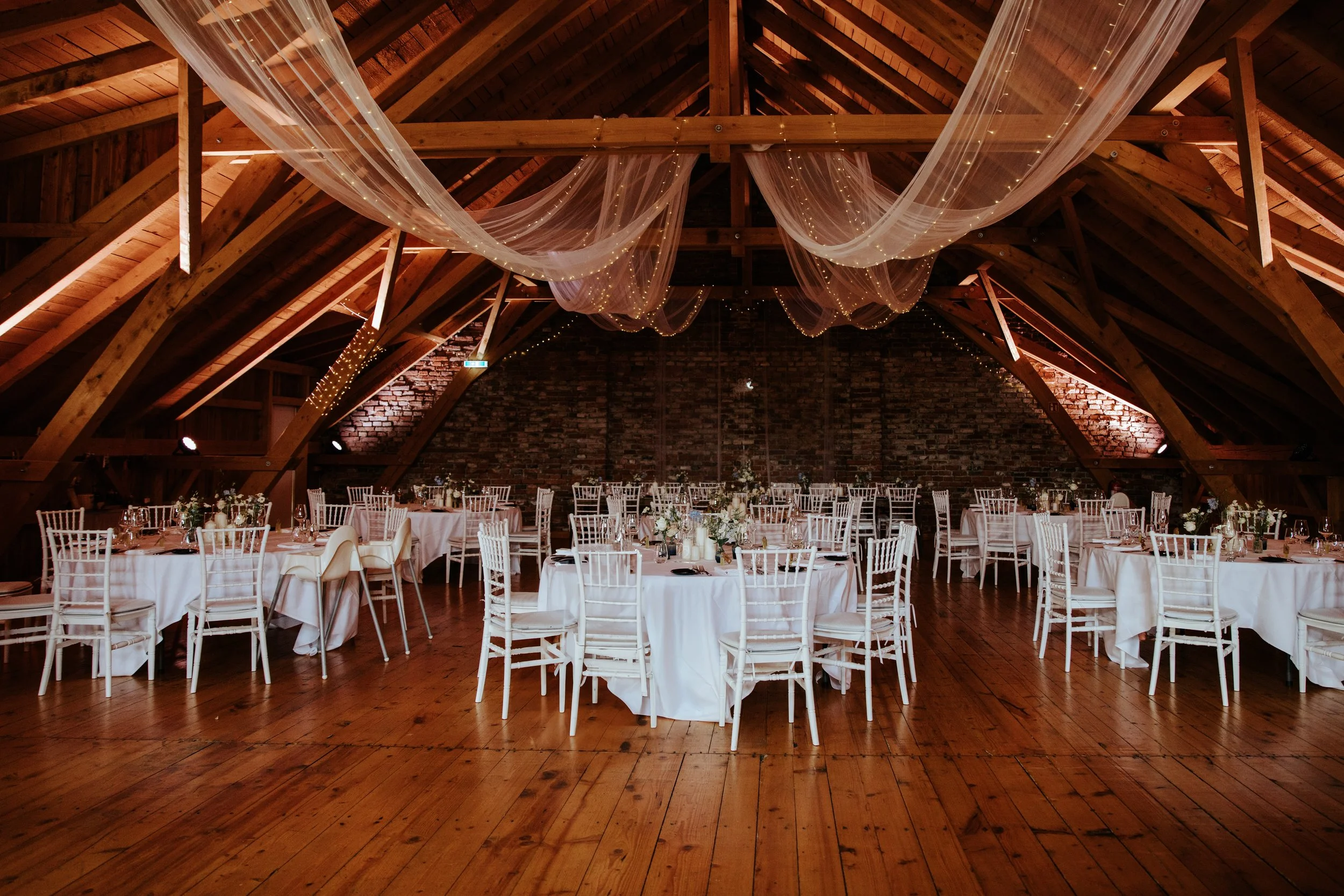 Eleganter Festsaal mit runden Tischen, weißen Stühlen und Dekorationen, in einem Raum mit Holzdecke und Backsteinwand, festliche Beleuchtung und Tüllvorhänge