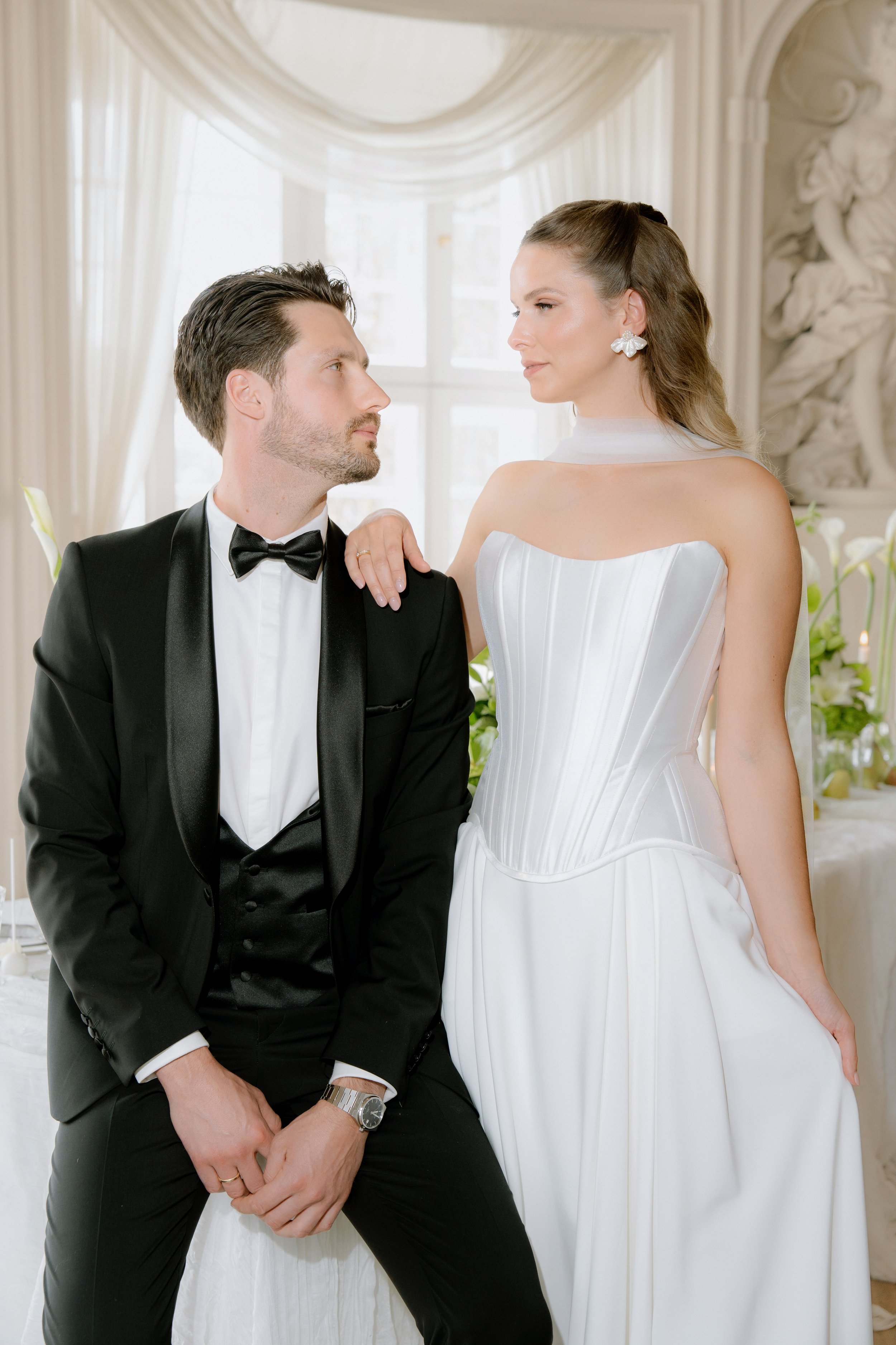 Ein Paar in Hochzeitskleidung; der Mann trägt einen schwarzen Anzug mit Fliege, die Frau ein weißes Kleid. Sie stehen in einem elegant dekorierten Raum mit Blumen.