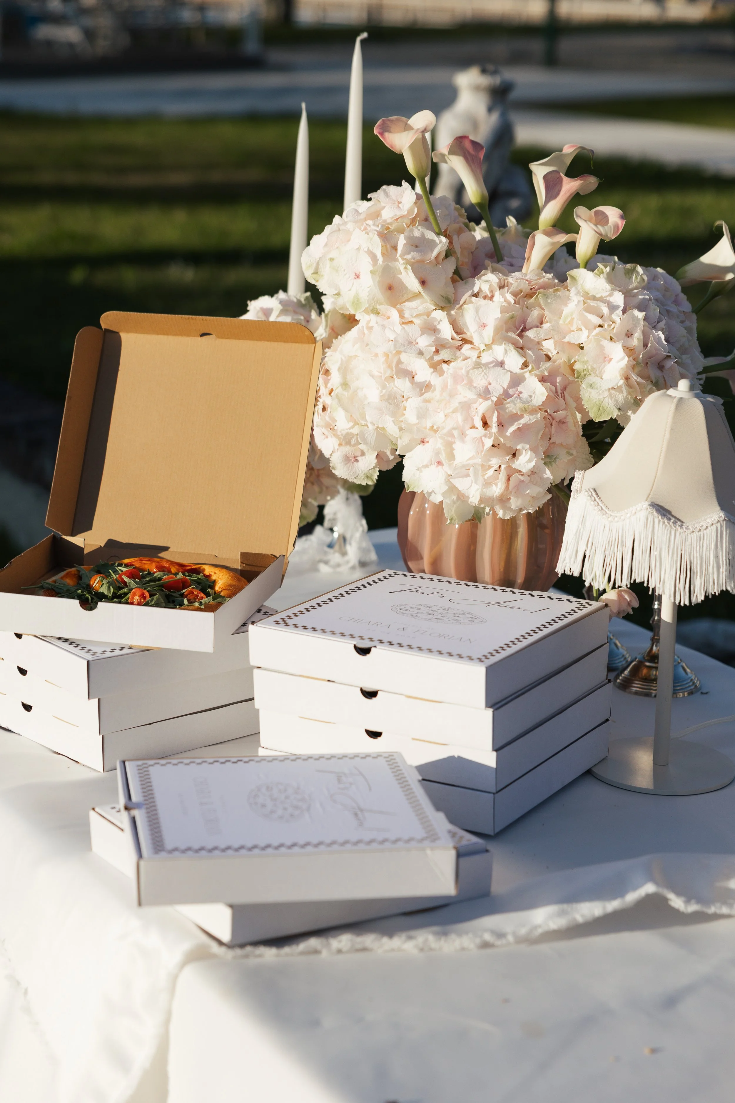 Auf einem Tisch befinden sich mehrere kleine weiße Geschenkboxen, ein Blumenstrauß aus weißen und pastellfarbenen Blumen, eine offene Pizzaschachtel mit Pizza, ein weißes Parfümfläschchen und ein kleiner Lampenschirm. Im Hintergrund ist eine grüne Wi