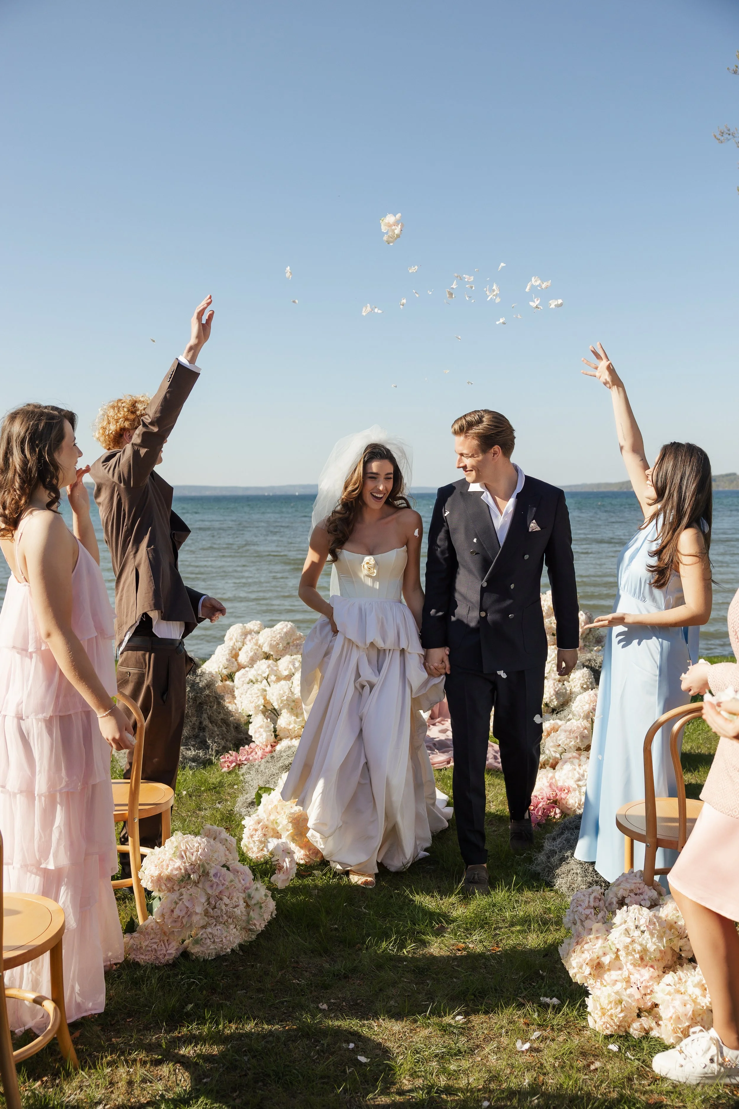 Ein Brautpaar und Gäste bei einer Strandhochzeit, Blumen, Wasser, blaue Himmel.