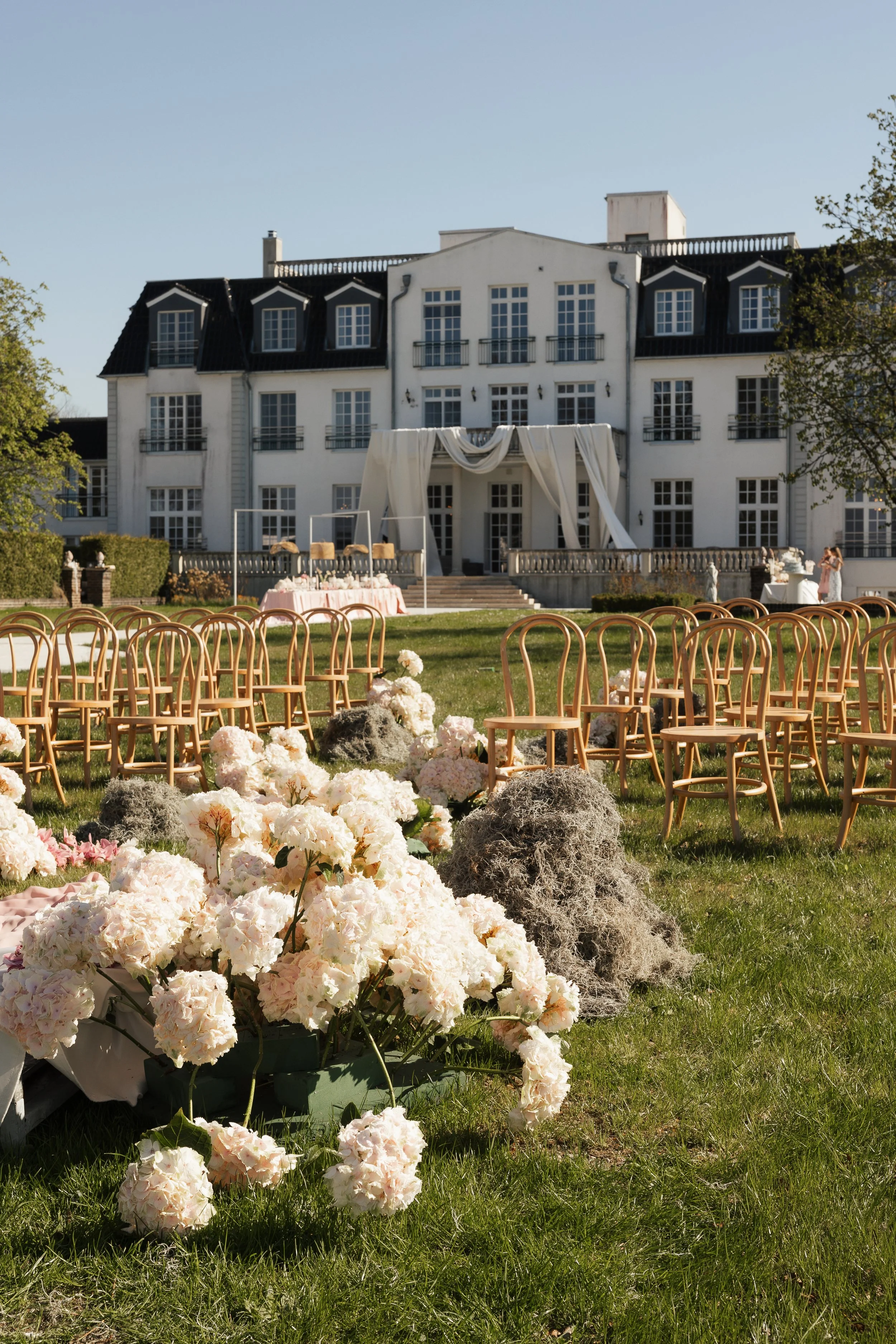Ein großes weißes Herrenhaus mit mehreren Stockwerken und schwarzen Dächern ist im Hintergrund sichtbar. Im Vordergrund befindet sich eine Gartenfläche mit Stroh- und Holzkörben, blühenden weißen und rosa Blumen sowie Trockengewächsen. Es gibt auch S