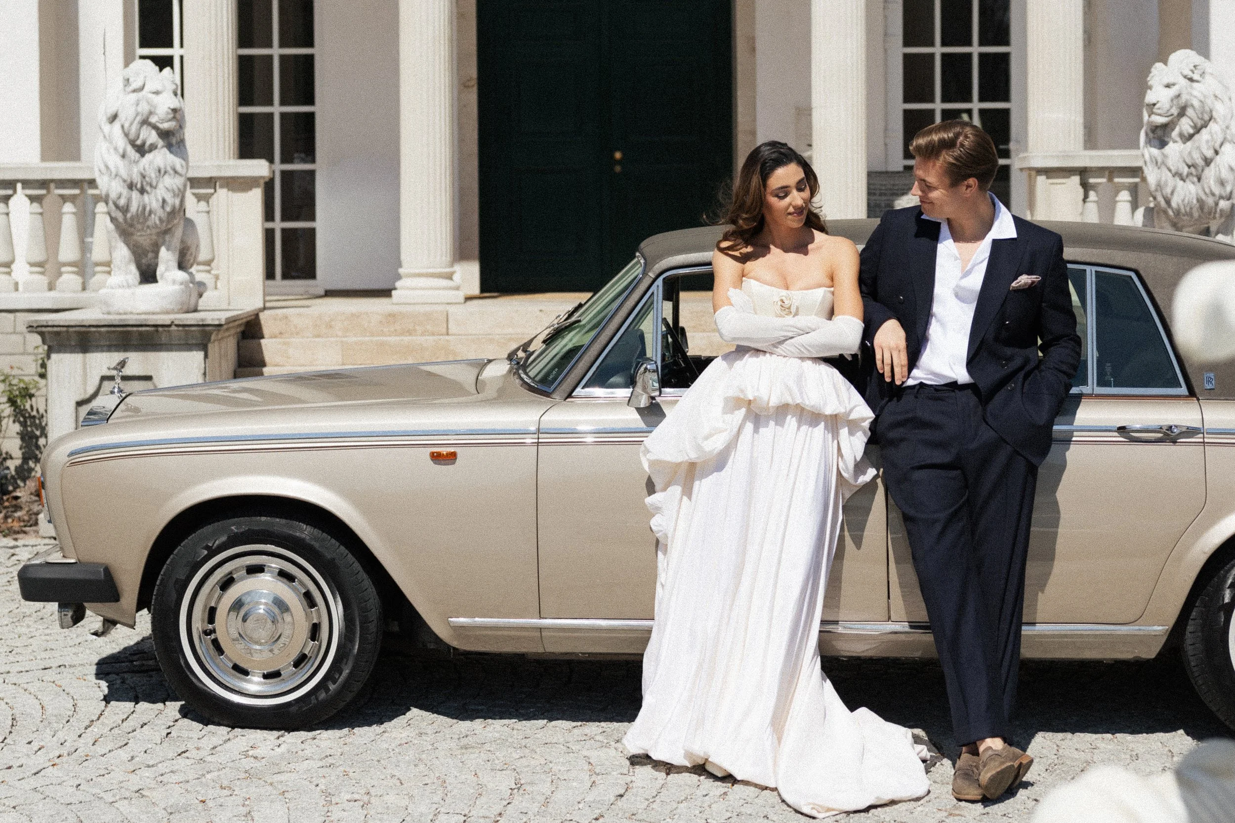 Ein Mann und eine Frau stehen vor einem klassischen Auto vor einem großen, weißen Haus mit Säulen. Das Mädchen trägt ein weißes Hochzeitskleid, der Mann einen dunkelblauen Anzug.