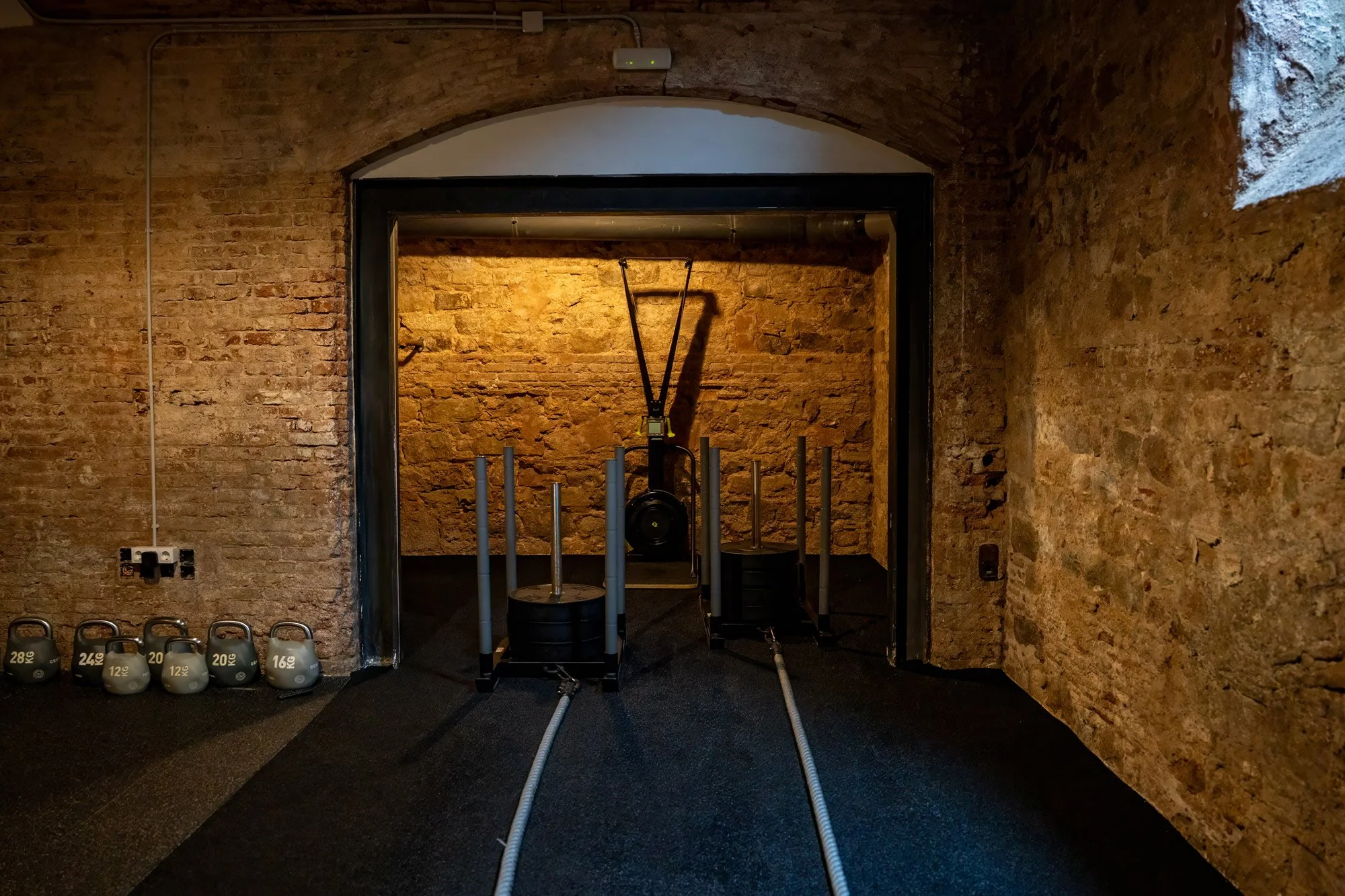Indoor gym space with black rubber flooring, stacked kettlebells on the left, and a black and yellow suspension training system in the center against a brick wall.