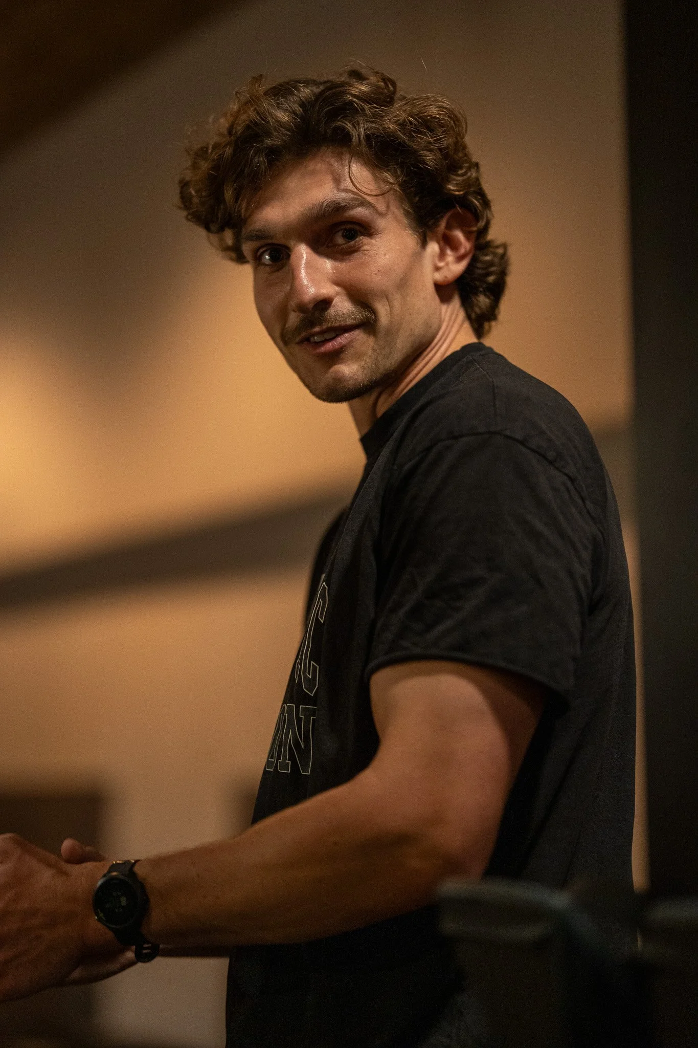 A young man with curly brown hair looking at the camera, wearing a black t-shirt and a black watch, standing in an indoor setting with warm lighting.