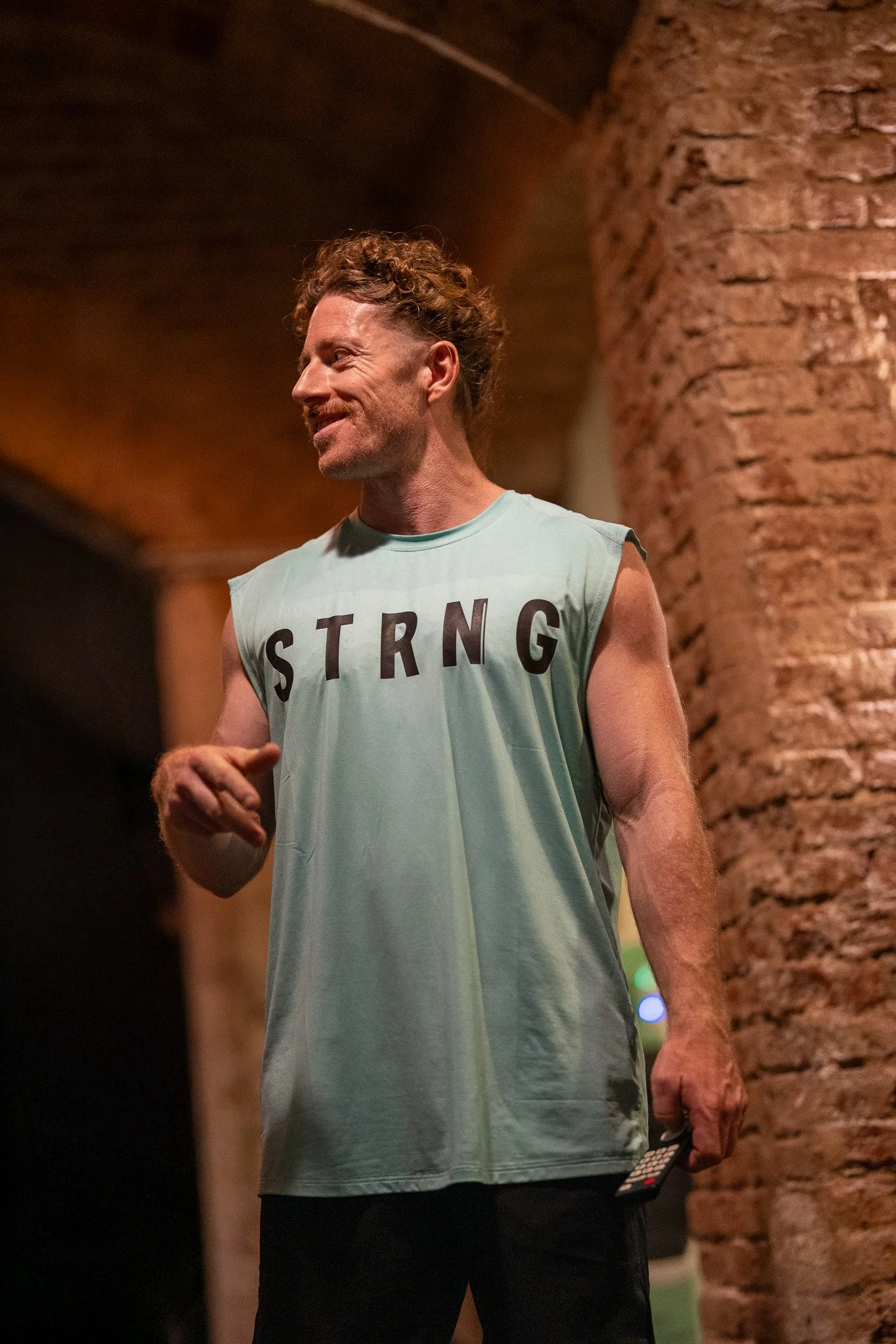 A man with curly hair and a beard smiling, wearing a sleeveless light-colored shirt with the word 'STRONG' written across it, standing indoors near a brick wall.