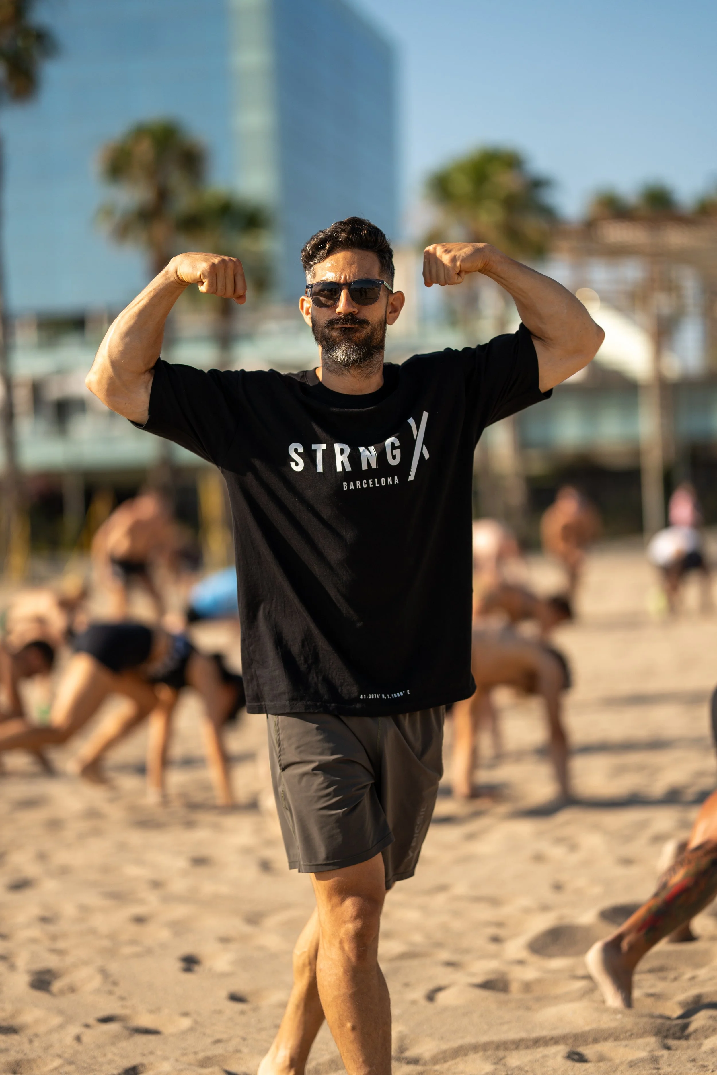 A man with dark hair, beard, sunglasses, and a black t-shirt that says 'STRONG Barcelona' is flexing his biceps while standing on the beach. In the background, people are exercising in a yoga or workout class on the sand.