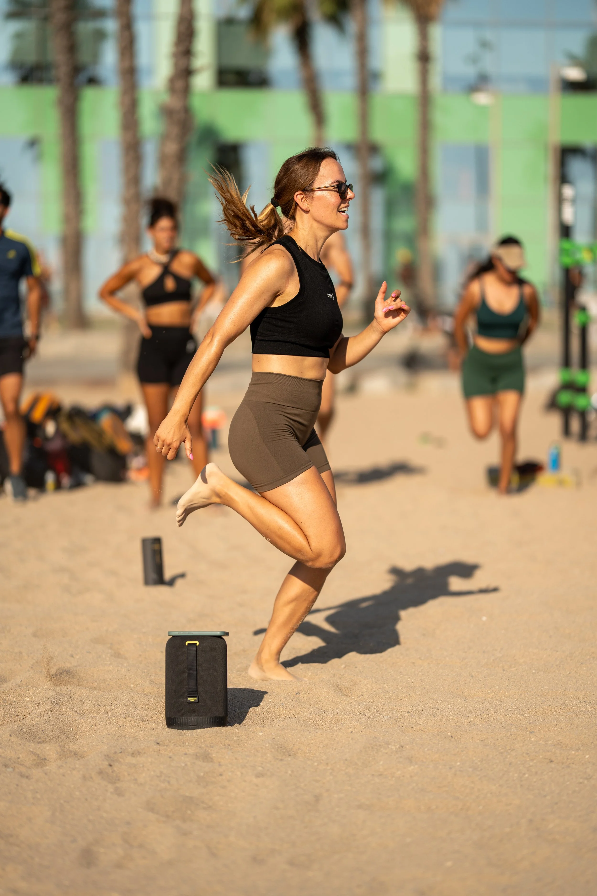 A woman with long hair jumping over a small black speaker on a sandy beach during a workout or fitness class, with others exercising in the background.