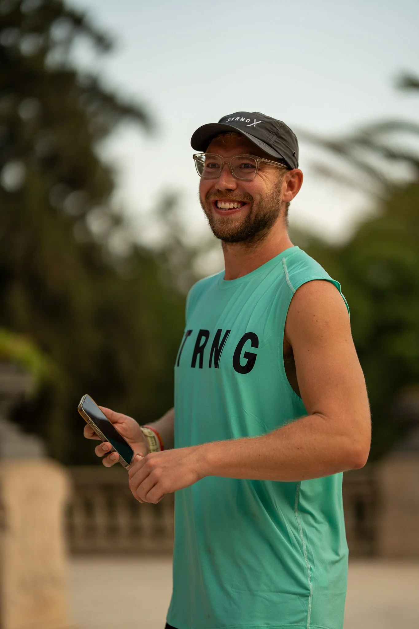 A smiling man wearing glasses, a black cap with white text, and a sleeveless teal running shirt with the word 'STRONG' printed on it, holding a smartphone in his right hand outdoors.
