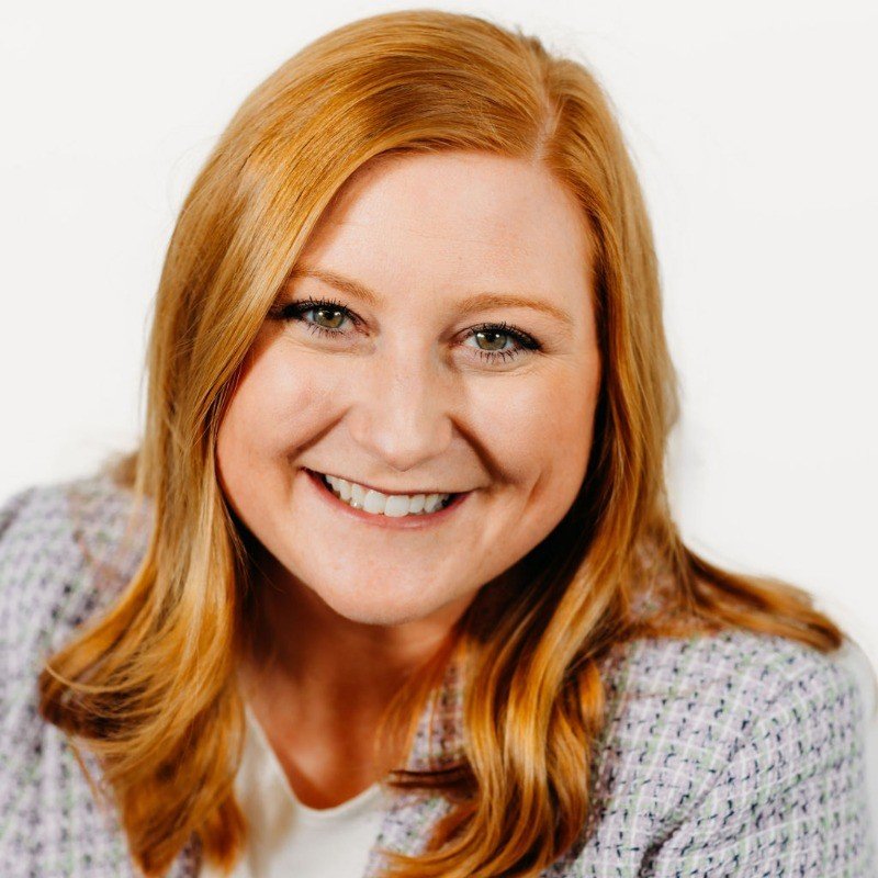 Headshot of a smiling woman with long red hair, blue eyes, wearing a checked blazer and white top, against a white background.